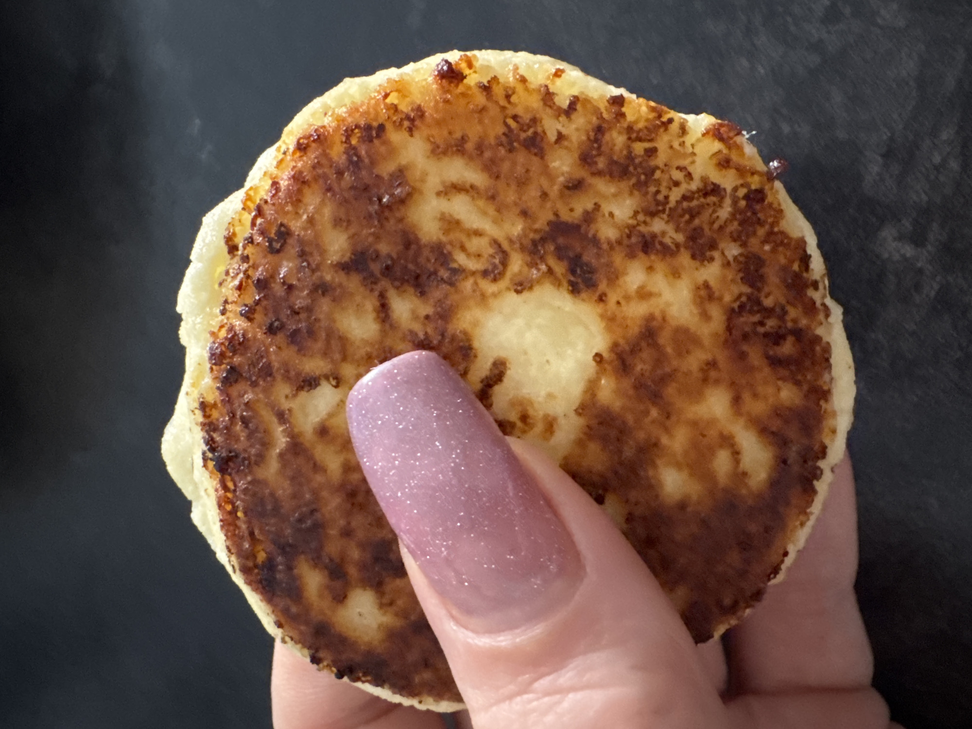 A close-up, high-angle shot features a hand holding a round, golden-brown food item against a dark, out-of-focus background. The food, possibly a pancake, fritter, or cheese curd pancake (syrnyk), displays a crispy, well-cooked surface with varying shades of brown and lighter, more yellow areas. The surface texture suggests it was pan-fried.
A human thumb, with a long, oval-shaped nail, holds the food item from the front. The nail is painted with a glittery, translucent light pink or lavender polish. The skin of the hand appears light-toned, with visible lines and creases on the thumb and the beginnings of other fingers in the lower right corner. The background is uniformly dark and appears smooth, providing a stark contrast to the brightly lit food and hand. There are no visible location cues within the image to suggest Poznan, Poland. No text is visible in the image.