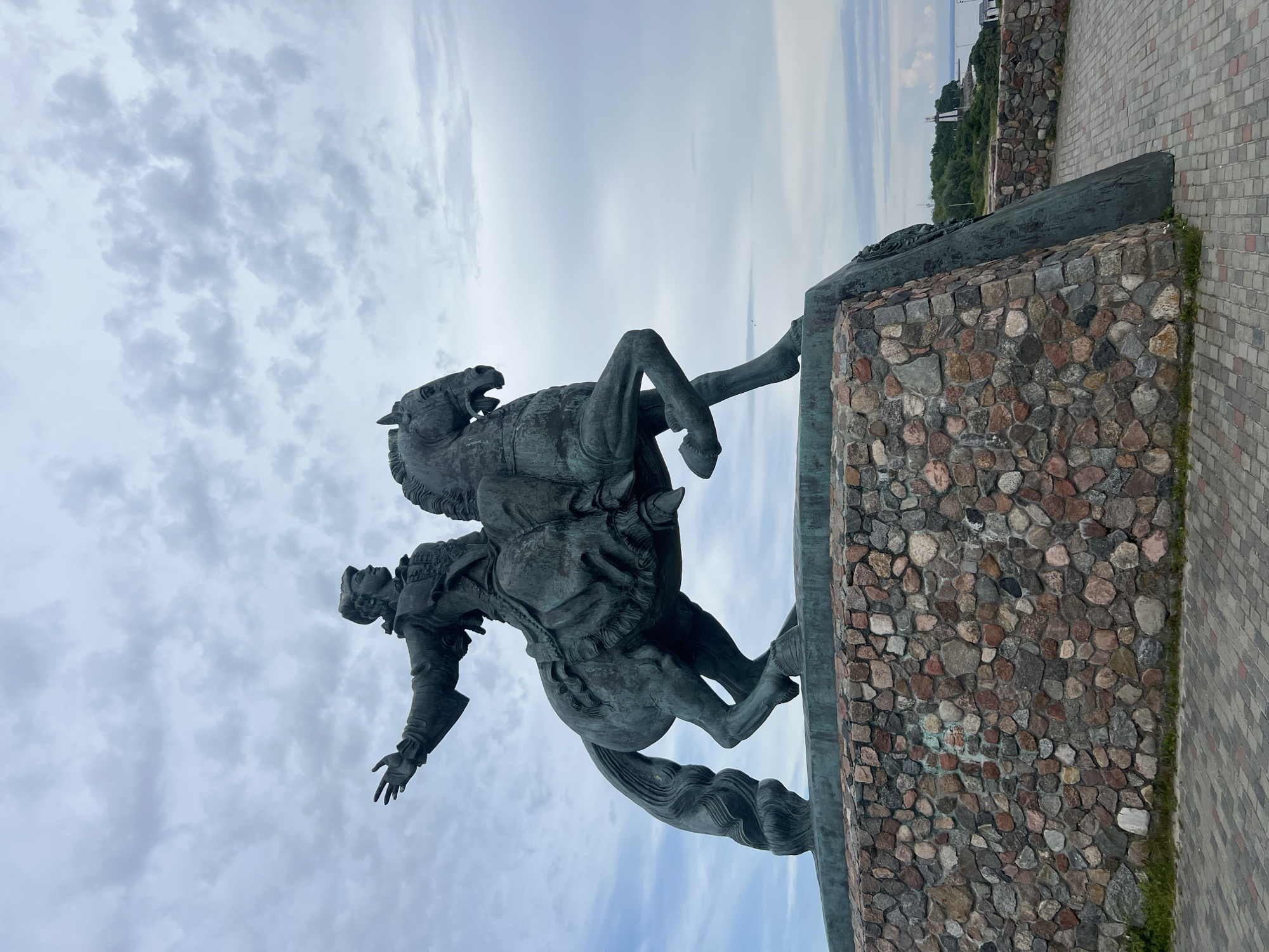 An equestrian statue of Peter the Great stands on a stone base in Baltiysk, Russia. The statue is made of bronze and depicts Peter the Great in a historical costume, pointing forward with his left hand. The horse is rearing up on its hind legs. The base of the statue is made of rough-hewn stones. In the background, there is a body of water, a lighthouse, and some trees. The sky is overcast with clouds.