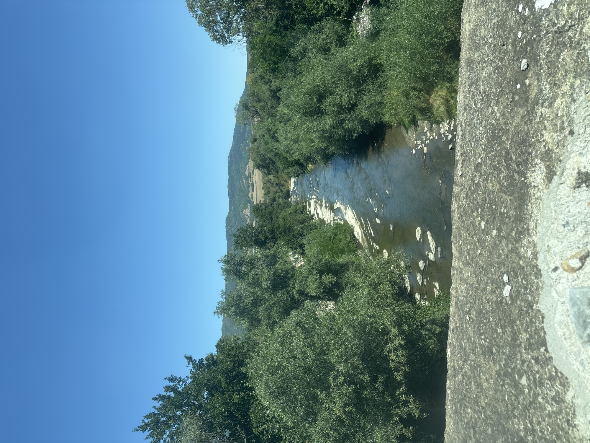 The image depicts a scenic view of a river flowing through a lush, green landscape under a clear blue sky. In the foreground, a textured, grey concrete structure, possibly a bridge or wall, occupies the bottom of the frame. Beyond this, a river meanders through the scene, its water reflecting the sky and surrounded by dense foliage. The riverbed appears to have some rocky outcrops and areas where the water is shallow. Trees line both sides of the river, creating a verdant corridor that extends into the distance. In the background, rolling hills covered in vegetation are visible under the expansive sky. The overall impression is one of natural beauty and tranquility. While the image is identified as being in Vidrare, Bulgaria, there are no specific landmarks or details that pinpoint this exact location beyond the general description. There are no people or animals visible in the image.