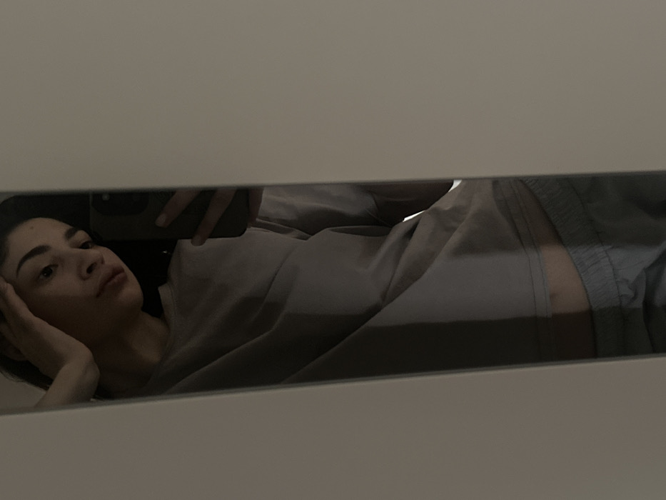 A female individual is depicted in a dimly lit mirror selfie, taken indoors in Yekaterinburg, Russia. The subject, with dark hair and prominent eyebrows, appears to be an adult in her twenties or thirties, captured from the chest to the upper thighs. She is wearing a light grey or beige short-sleeved t-shirt and light grey athletic shorts, with a visible section of her lower abdomen. Her right hand is positioned against her forehead, while her left hand holds a dark-colored smartphone, which is actively taking the mirror image. The visible environment immediately around the mirror consists of a plain, light-toned vertical surface, likely a wall or door frame.