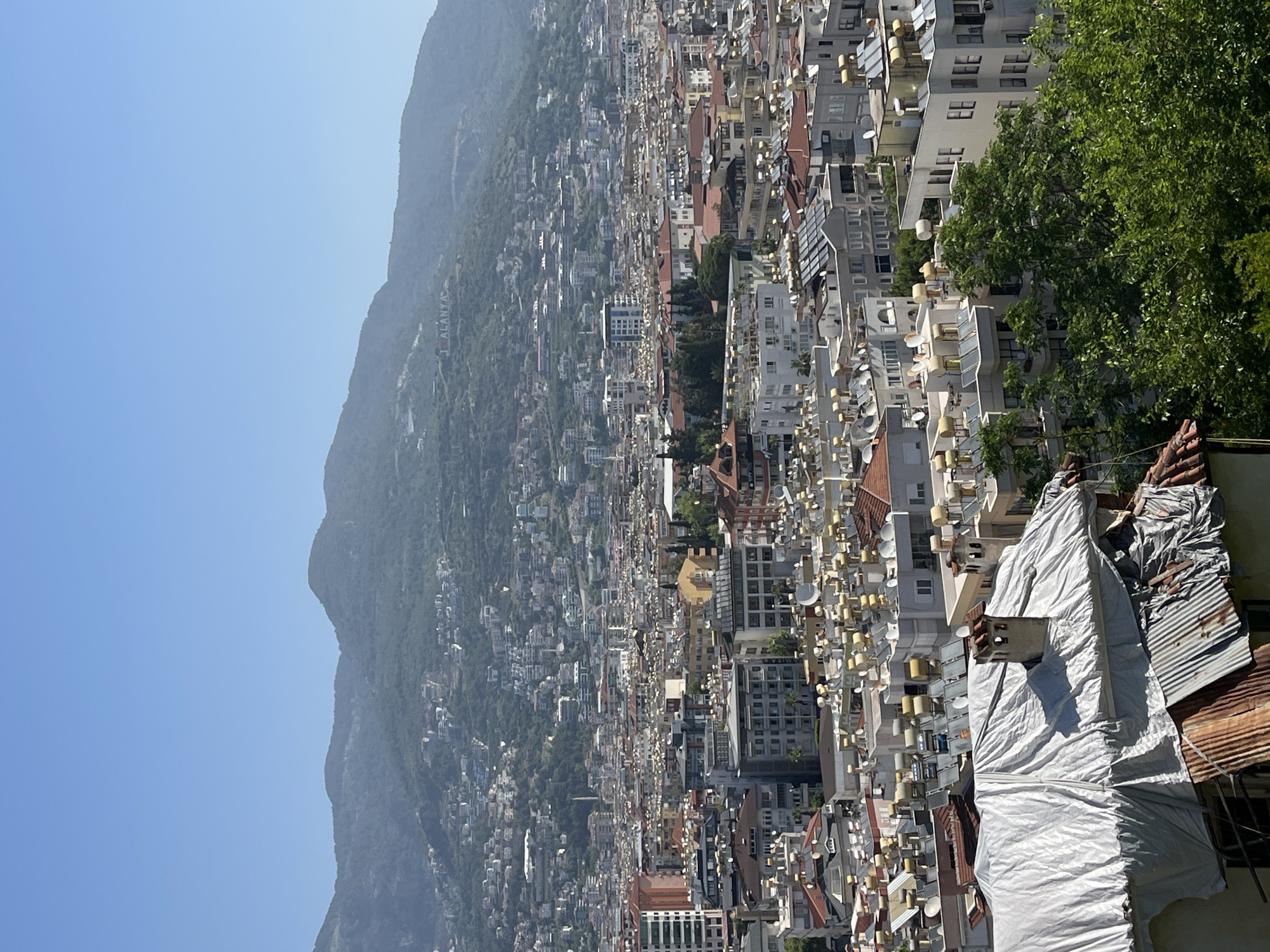 The image shows a high-angle view of Alanya, Turkey, likely taken from a vantage point overlooking the city and the adjacent mountain.
Environment: The scene is set in a densely populated urban area stretching from the base of a large, green, tree-covered mountain towards the coast. The sky is clear and blue.
Objects: Numerous buildings of varying heights and architectural styles dominate the scene, mostly white or light-colored. Many buildings have satellite dishes mounted on their roofs. The mountain is heavily wooded, and there are buildings nestled within the trees along the lower slopes. In the foreground, there's a structure covered with a silver-colored tarp and some weathered corrugated metal roofing.
Location: The presence of the mountain and the dense urban layout suggest that this is Alanya, Turkey. The lettering visible on the mountain "I <3 ALANYA" further confirms the location.
People/Actions/Interactions: No people are immediately visible in the image. There are no specific actions or interactions that can be identified.