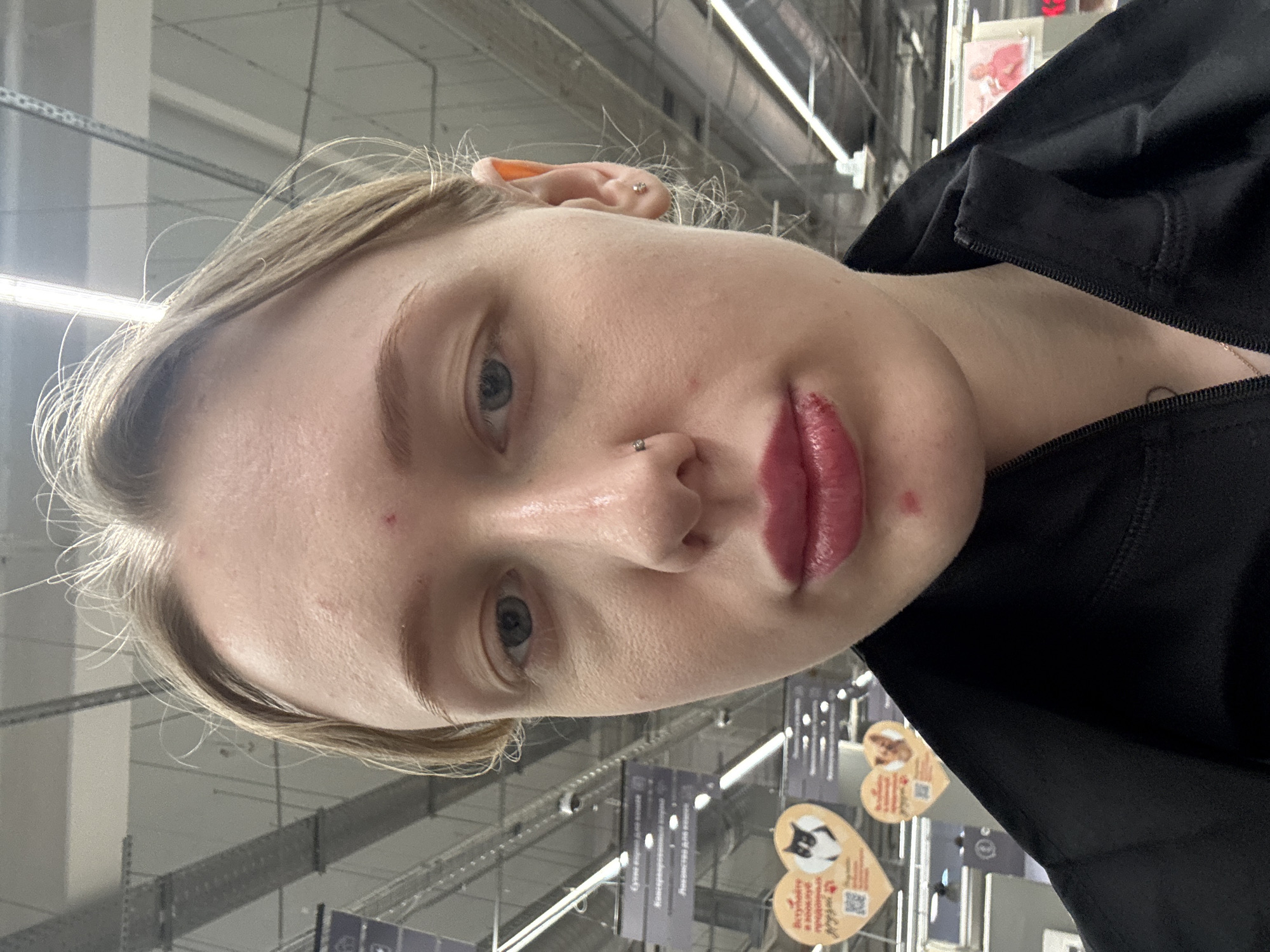 A close-up, rotated shot of a fair-skinned young adult with blonde hair. The individual has light blue eyes, a piercing in their left nostril, and earrings. They are wearing a black jacket. The background is blurred, showing elements of what appears to be a retail environment, including signage and overhead lighting. Based on the presence of signage written in Russian, and the prompt's mention of Rostov-on-Don, it is likely that this image was taken in a store in Rostov-on-Don, Russia. The focus is on the person's face and immediate surroundings, giving little information on specific actions or interactions.