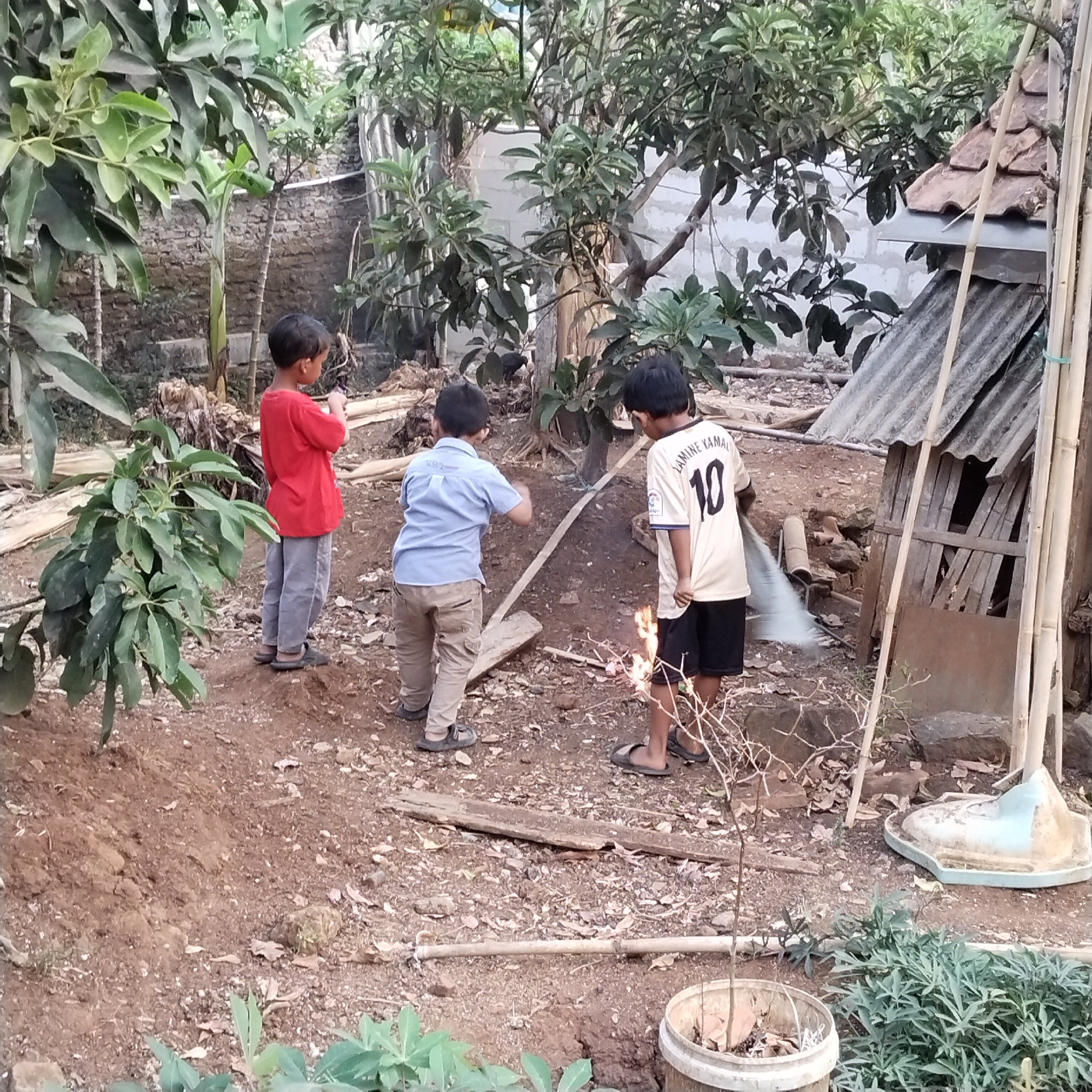 The image depicts an outdoor scene in the city of Pamulihan, Indonesia, likely a rural or semi-rural area given the natural vegetation and rustic structures. Three young boys are visible. The boy on the left wears a red t-shirt and grey pants. The boy in the middle wears a light blue button-down shirt and khaki cargo pants. The boy on the right is wearing a yellow and white jersey with the number "10" on the back and black shorts. He appears to be holding a broom or similar tool and sweeping near a small fire or burning debris. The ground is composed of dirt and scattered leaves. In the background, there are trees, a brick wall, and a small, crudely built structure with a corrugated metal roof and tiled accents. Several bamboo poles are also present, leaning against the structure and standing upright. The overall impression is of a casual, outdoor setting, possibly a backyard or open lot where children are engaged in activities.