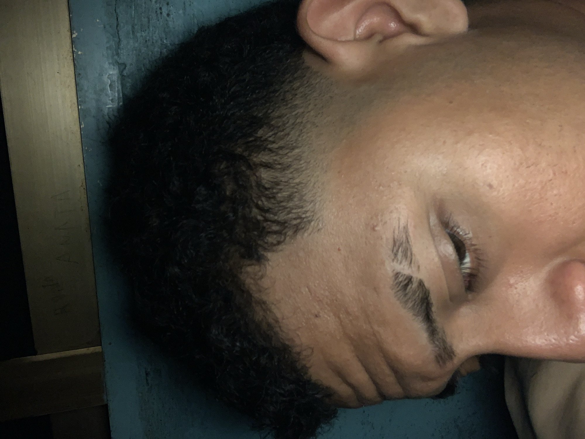 The image displays a close-up, side-profile view of a person's head, appearing to be a male, resting against a surface. The person has short, dark, curly hair with a clear fade haircut on the side of their head. Their skin is light brown or tan, with visible pores and some creases on the face. The eyes are closed or partially closed, suggesting the person is asleep or deeply resting. A notable detail is a distinct gap or cut in one of the eyebrows, possibly a scar or a stylistic feature.
The setting appears to be indoors, with the person's head positioned against a light blue or teal surface, which could be a bed, pillow, or wall. To the left, a light-colored wooden beam or frame is visible, extending vertically. Faint, dark text is etched or written on this wooden surface, partially decipherable as "RADO ANNA" or "RADO ANA". The lighting is dim, casting soft shadows and contributing to a subdued atmosphere. The image captures a moment of quiet repose in what appears to be Santa Ana, Venezuela.