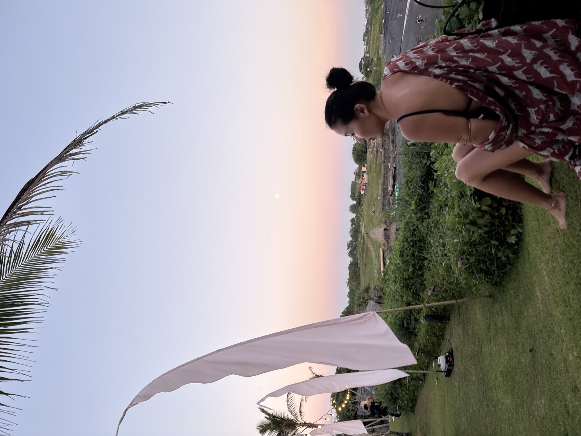 The image shows a woman crouching and looking down in an outdoor setting. She has her hair in a bun, wears a dark top, a patterned burgundy robe, and gold bracelets. The background suggests a tropical location, possibly a cliffside, with lush greenery and thatched roof structures. Several white flags are visible, along with palm trees and string lights. In the distance, there are structures that may be part of a village or resort, and a rainbow is visible in the sky. The location is likely in Kediri, Indonesia, given the environmental cues and architectural styles present.