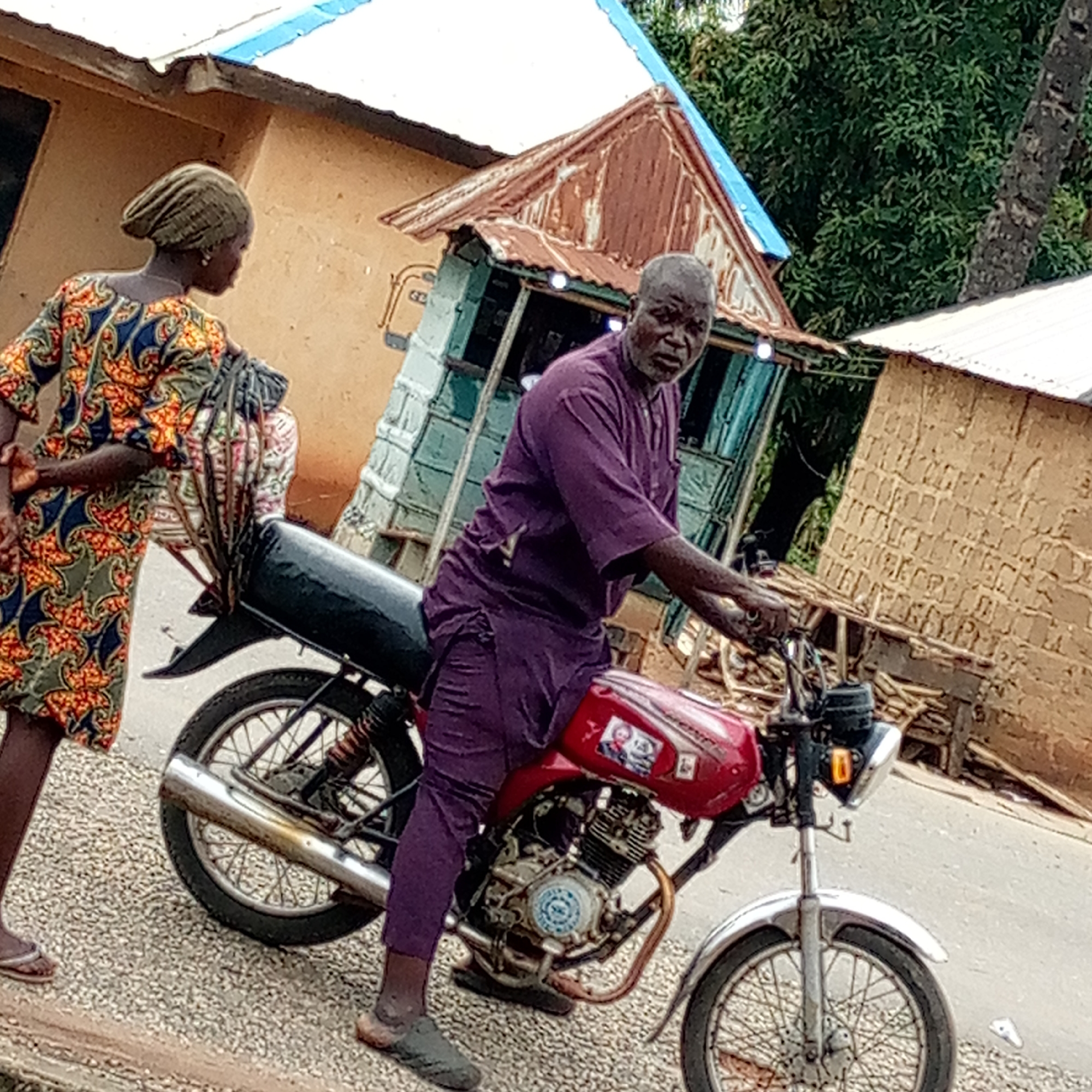 In Mararaba, Nigeria, an elderly man in a purple outfit sits astride a red and black motorcycle on a gravel road. The motorcycle has a silver front fender and a sticker on the gas tank. A woman in a colorful patterned dress and headwrap stands beside the motorcycle, holding a bundle of items. Behind them are several small, worn buildings with corrugated metal roofs. Debris is scattered near the buildings.