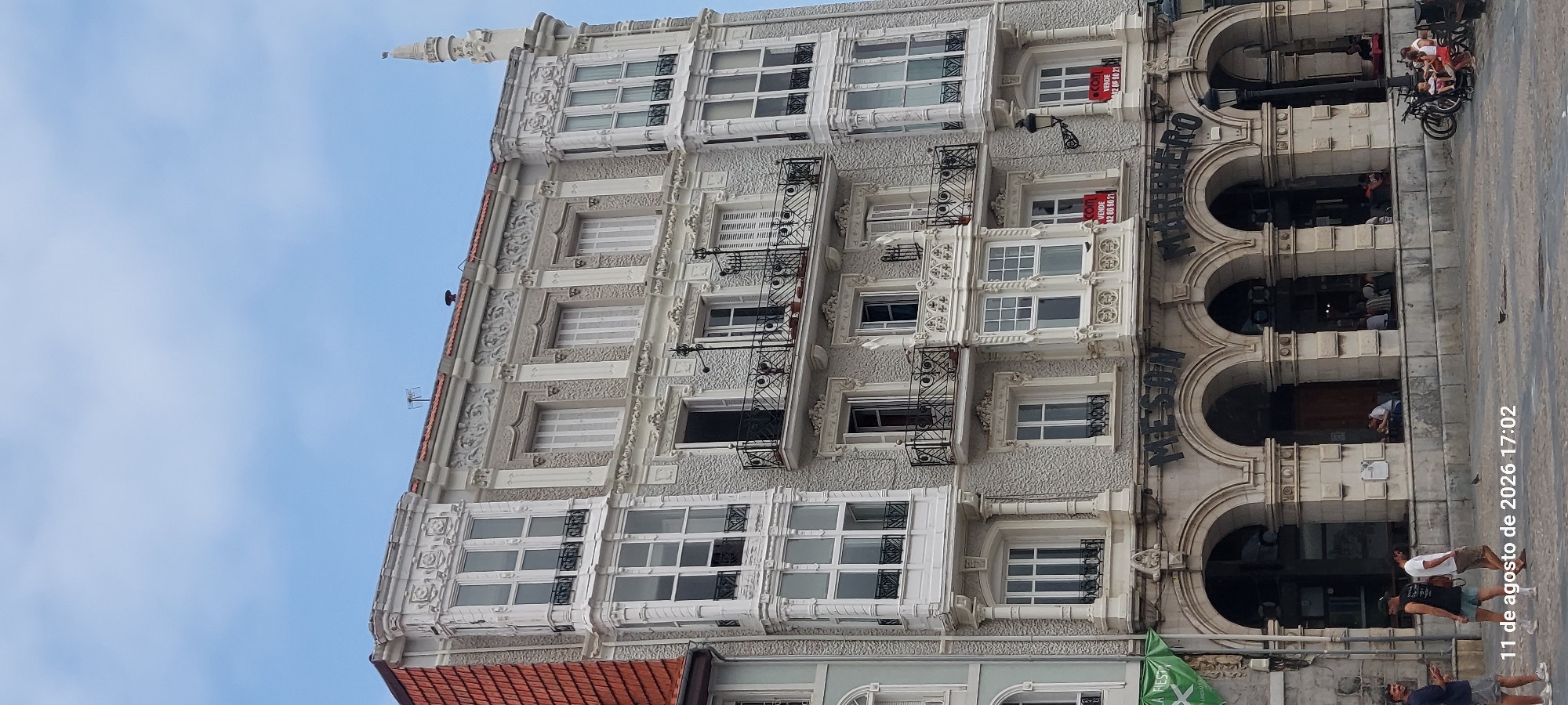 The image shows a multi-story building in Castro-Urdiales, Spain. The building has a decorative facade with ornate white trim, balconies with black wrought-iron railings, and large windows. The ground floor features arched entrances that appear to lead into a restaurant or bar named "MESON MARINERO." Several people are visible in the foreground, with some sitting outside the establishment and others walking on the pavement. There are also bicycles and a stroller parked nearby. The sky is partly cloudy. The timestamp "11 de agosto de 2026 17:02" is visible in the bottom left corner.