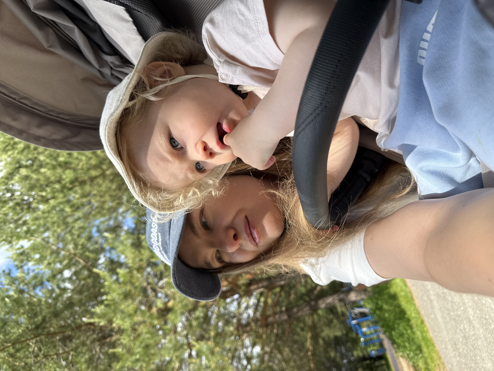 This outdoor selfie, taken in Dobrograd, Russia, captures a woman and a young child in what appears to be a stroller or pram on a sunny day. The image is rotated 90 degrees clockwise from its natural orientation.
The subjects are positioned from bottom to top of the frame. At the bottom, a woman with long blonde hair wears a white t-shirt and a dark blue baseball cap, which has partially visible text including "COAST". She looks towards the camera with a slight smile. Above her is a young child, likely a toddler or baby, with blonde hair and blue eyes. The child is nestled in the stroller, dressed in a light-colored top, and has their fingers in their mouth, looking directly at the camera with a curious expression. The dark frame of the stroller is visible around the child.
The background features lush green foliage from trees and patches of blue sky, indicating bright daylight. In the lower right, a paved path and a blurry blue structure, possibly a playground feature or fence, are visible. Next to the child, a light blue piece of fabric with the brand name "PUMA" printed in white letters is clearly visible. The overall scene suggests a pleasant, casual moment outdoors between a parent and child.