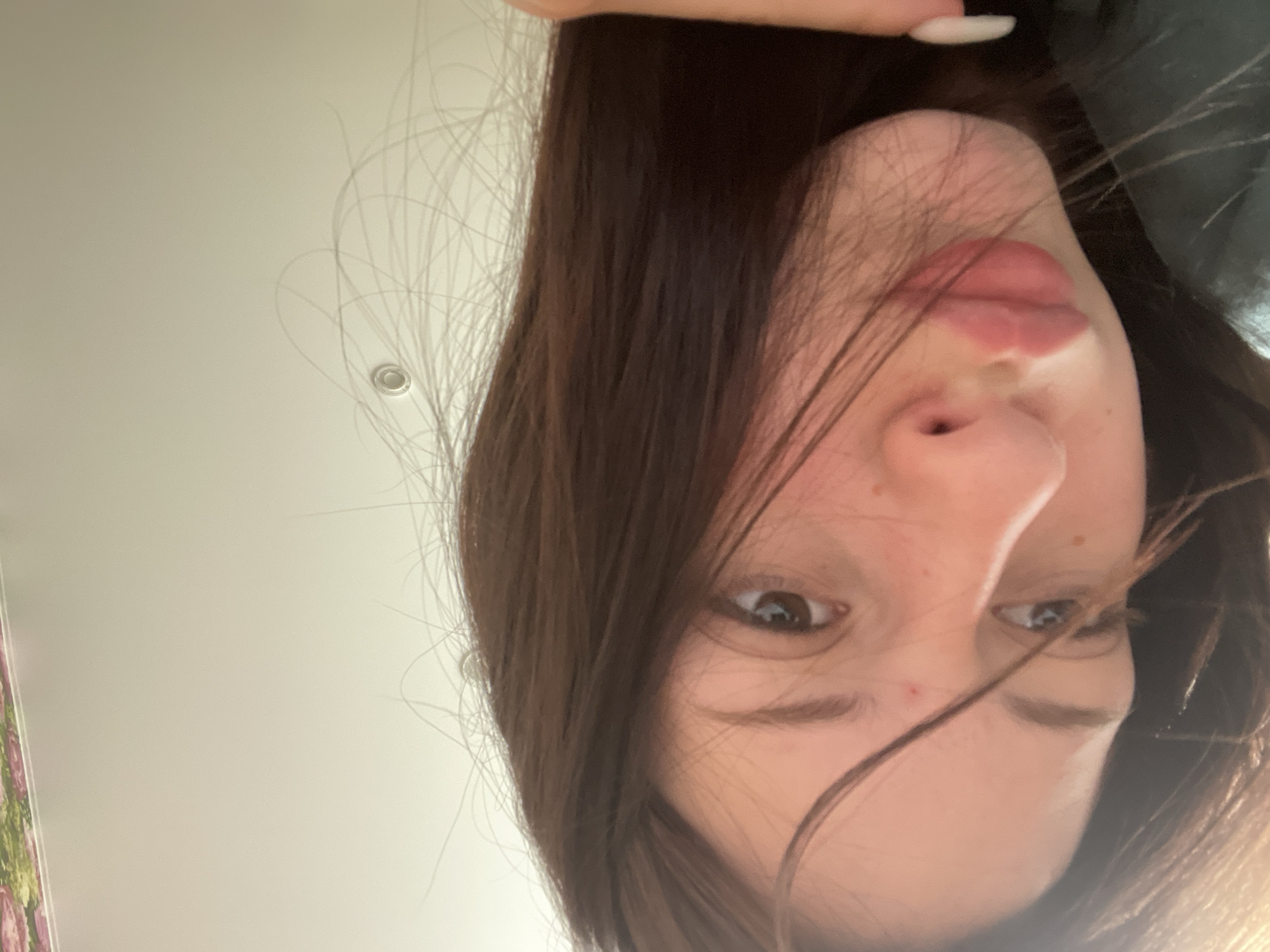 The image displays a close-up, low-angle perspective of a person, likely a young woman, photographed from below. The subject's head is tilted downwards, and her face is looking upwards towards the camera, with the image rotated ninety degrees counter-clockwise, making her appear on the right side of the frame. Her long, dark brown hair falls around her face, partially obscuring one eye. One dark eye is clearly visible, with a contemplative or neutral expression, looking towards the left of the frame. Her lips are slightly parted, and a portion of her nose and chin are visible. A small red mark is present on her cheek or forehead. The tip of a finger with a white manicured nail is visible at the very top edge, suggesting she is holding the camera.
The background is predominantly a plain, light-colored surface, appearing to be a ceiling or upper wall, indicating an indoor setting. A small, circular, metallic-rimmed object, possibly a light fixture or smoke detector, is visible on this surface towards the upper-middle left. To the far left, a blurred edge of a patterned fabric, possibly floral with pink and green hues, can be seen. The lighting is bright and even. The activity is a self-portrait taken from an unconventional, upward-looking angle. There is no visible text. The location is Медведево, Russia.