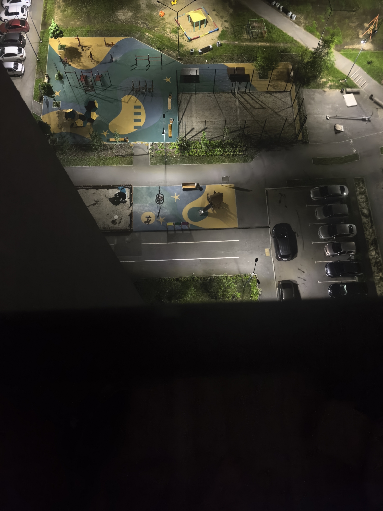 This is an aerial nighttime photograph of an urban playground and parking lot. The playground is brightly lit with colorful surfaces, featuring swings, climbing structures, and a yellow playhouse with a sandpit. Adjacent to the playground is a paved parking area with several cars parked in rows. The scene is illuminated by artificial lights, casting long shadows across the ground. The overall impression is one of a quiet, orderly urban environment at night. The time of day is clearly night due to the artificial lighting and dark shadows. There are no visible people or animals in the scene, and the weather appears clear. The setting appears to be a residential area, indicated by the presence of a playground and parking lot. No text is visible in the image.