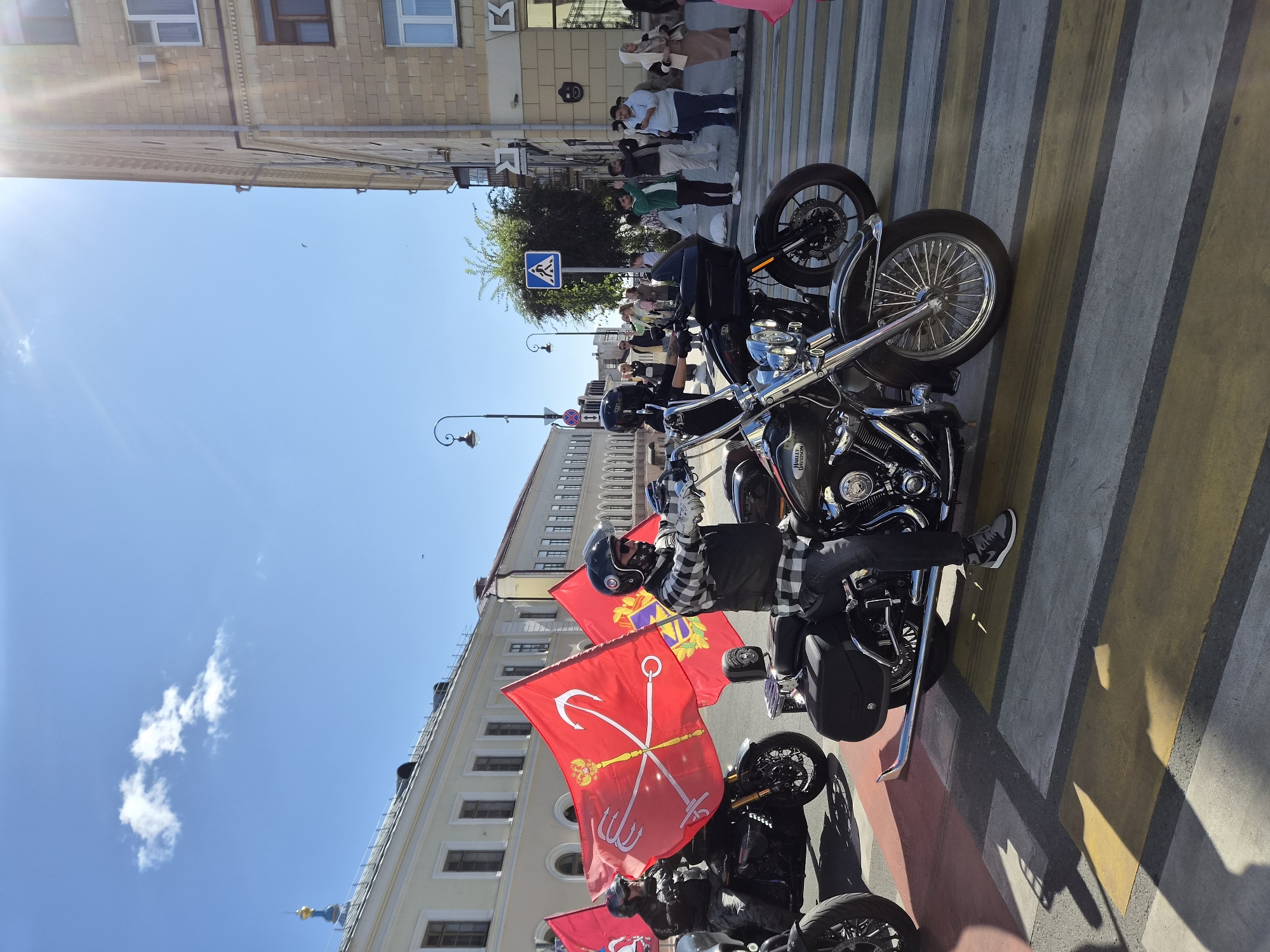 In Kazan, Russia, a group of motorcyclists rides through a crosswalk on a sunny day. The lead rider, wearing a black helmet and a plaid shirt over a vest, straddles a black Harley-Davidson motorcycle. Two large red flags with anchors, tridents, and coats of arms are prominently displayed, attached to the motorcycles of the riders in the foreground and middle ground. Behind them, several other motorcycles and riders are visible, and a crowd of onlookers stands on the sidewalk to the right, some with their phones raised to take pictures. The background features buildings, lampposts, and a clear blue sky with a few wispy clouds. The scene appears to be part of a parade or event.