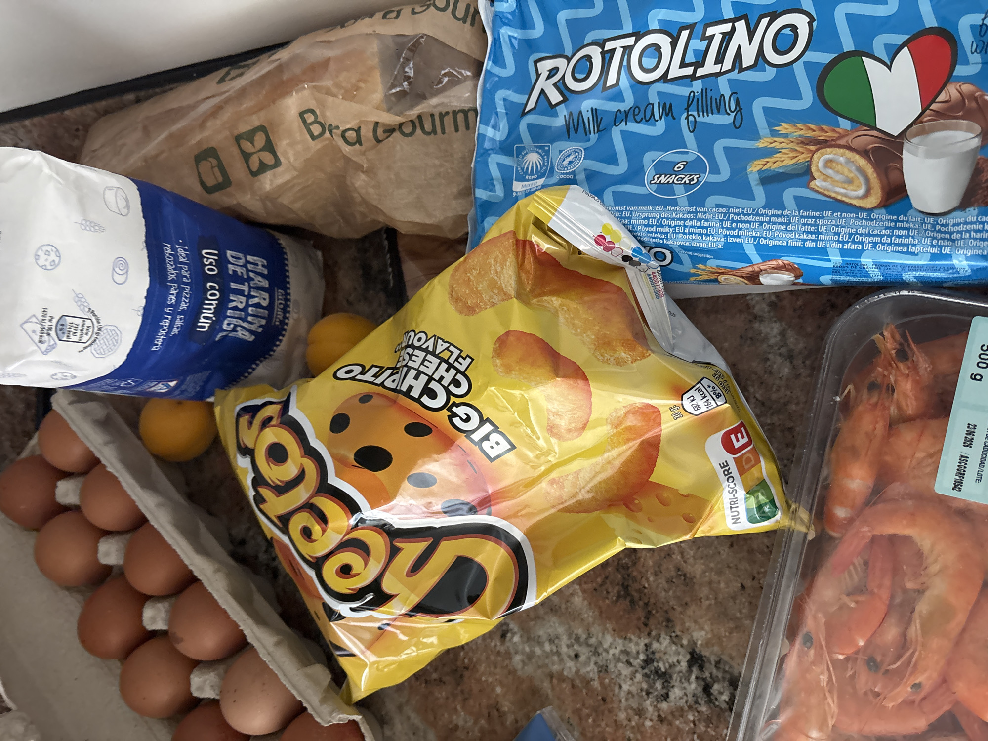 The image displays a collection of food items on a countertop, likely in a home kitchen in Orihuela, Spain. In the foreground, a package of "Harina de Trigo" (wheat flour) is visible, along with a bag of bread. To the left, a blue package of "Rotolino" snacks with "Milk cream filling" is present. Overlapping these items is a bright yellow bag of "Big Cheeto" cheese-flavored snacks. In the background, a container of cooked shrimp is partially visible. The countertop appears to be a speckled, granite-like material. The lighting suggests an indoor setting with overhead light sources. There are no people or discernible actions depicted in the scene.