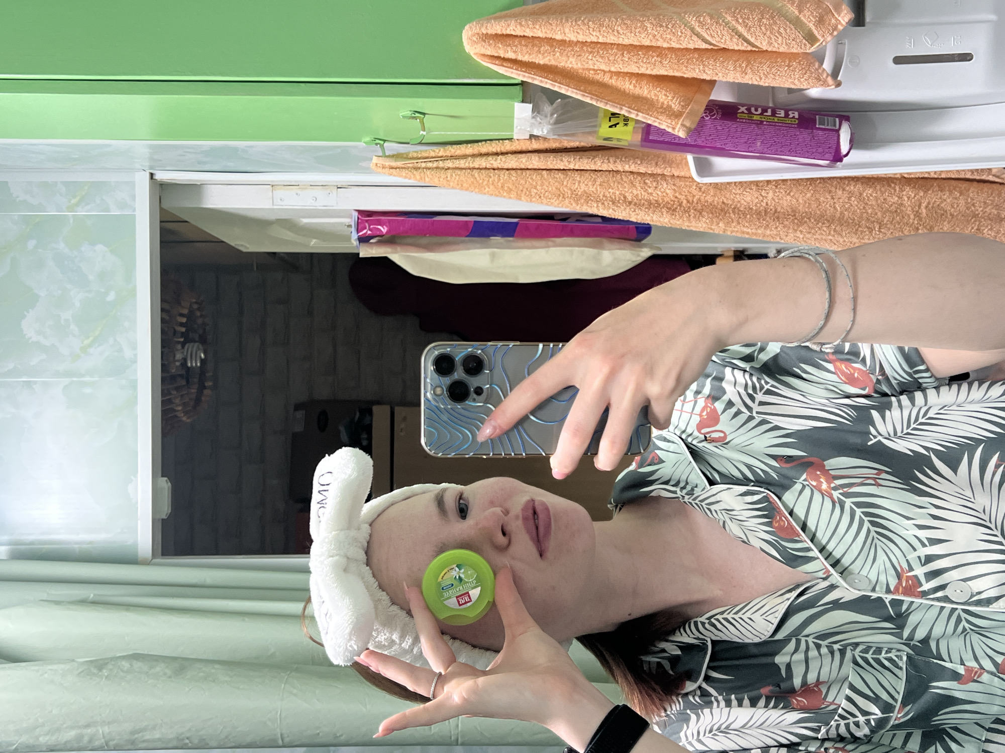 A young woman with fair skin and light brown hair, wearing a white terrycloth headband and pajamas with a palm leaf and flamingo pattern, holds a small green container in front of her left eye while taking a selfie in a mirror. The room appears to be a bathroom, with a green cabinet, towels, and a shower curtain visible. The mirror reflects part of the room and another door. The woman's right hand holds a smartphone. The overall lighting is artificial and appears to be from overhead.