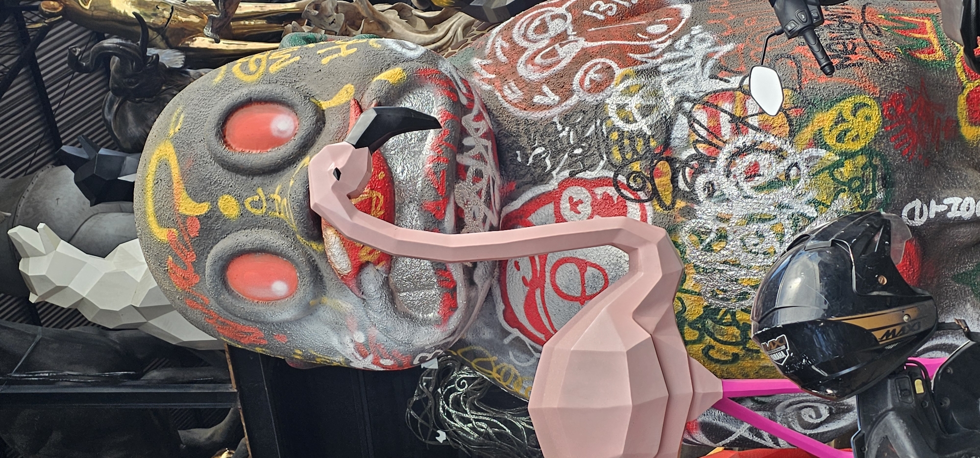 This photograph was taken in the Ubud District of Indonesia. It features a collection of eclectic objects and sculptures clustered together. The focal point is a large, stylized head with a textured gray surface, embellished with colorful graffiti and four prominent, red, circular "eyes." A geometric, light pink flamingo sculpture with an angular design is positioned in front of the head. A black motorcycle helmet with gold stripes and the "Maxi" label sits nearby, attached to a motorcycle. Other objects include a white geometric sculpture, a dark, sculpted animal figure, and a tangle of black cord or rope. The items appear to be displayed on a shelf or tiered arrangement.