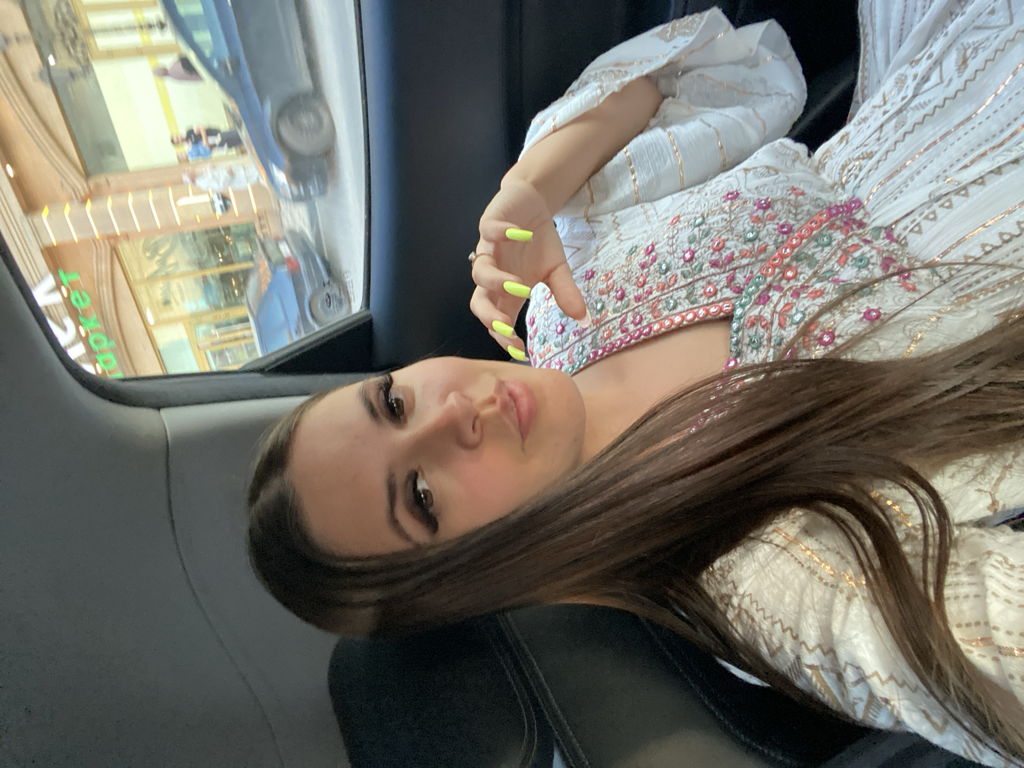 A young woman with long brown hair and bright yellow fingernails is seated in the back seat of a car. She is wearing a white dress with intricate embroidery in shades of pink, green, and gold. Her makeup includes dramatic winged eyeliner and full lashes. The car's interior is visible, with a dark leather seat. Through the car window, a street scene in Makhachkala, Russia, is depicted. Buildings with storefronts, including one with green lettering, line the street. Cars are parked and driving on the road, and a few pedestrians are visible on the sidewalk. The lighting suggests it is daytime.