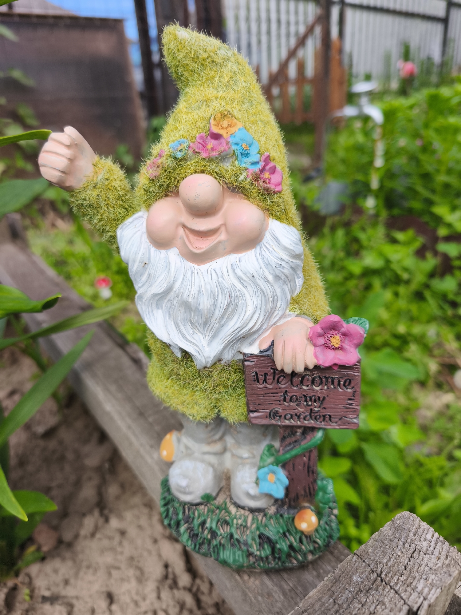 An outdoor, eye-level, medium shot focuses on a garden gnome standing on a wooden plank. The gnome, predominantly green and fuzzy, wears a pointed hat adorned with colorful plastic flowers. It has a rosy-cheeked, smiling face, a white beard, and is dressed in white pants and boots. The gnome is holding a small wooden sign that reads "Welcome to my Garden," decorated with a pink flower and a blue flower. The background features lush green foliage, a wooden fence, and a glimpse of a blue sky. The gnome is positioned in a garden setting.