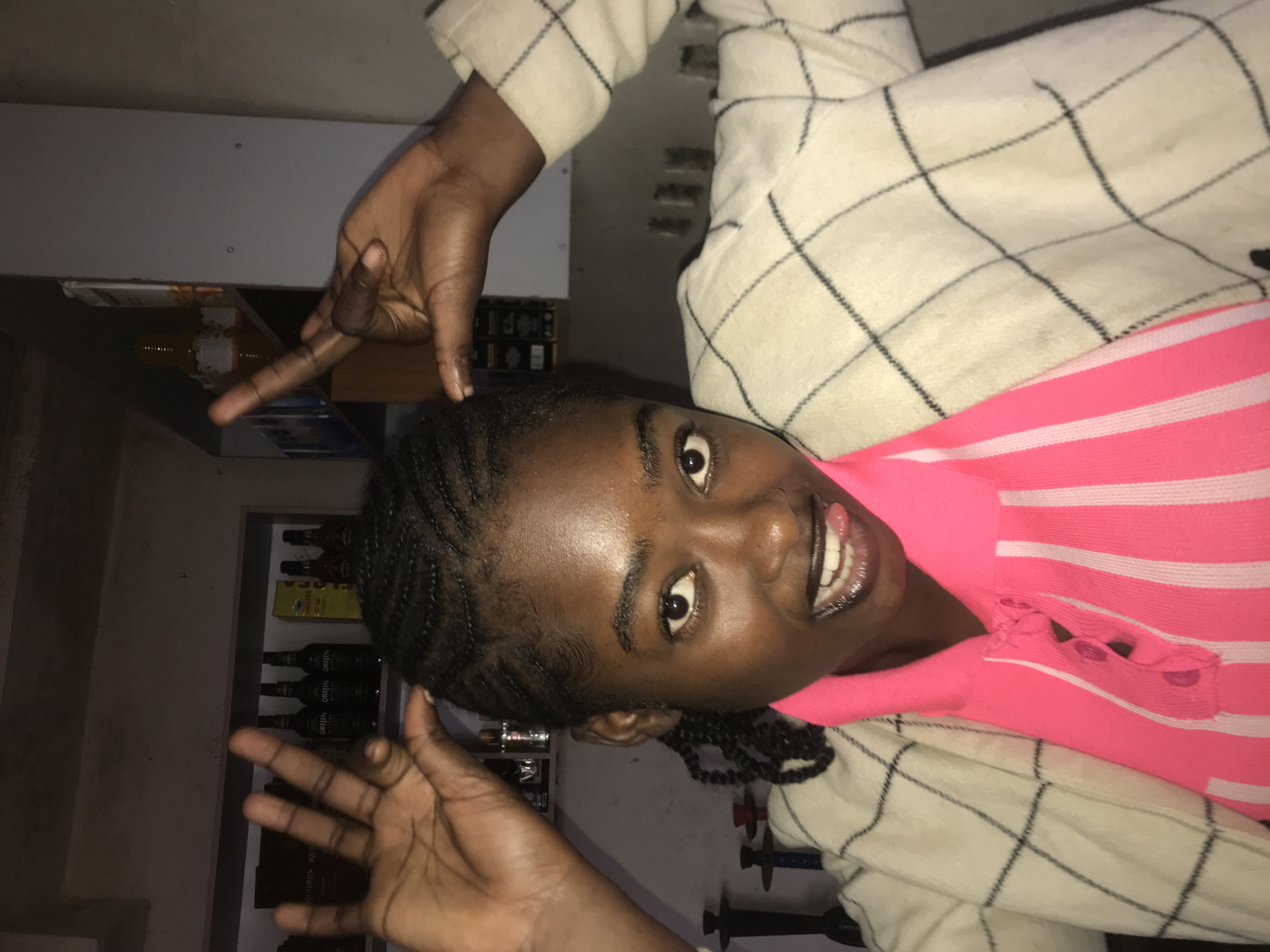 The image depicts a young woman posing indoors in the city of New Stadium, Nigeria. The subject, with a medium-dark skin tone and braided hair, is looking directly at the camera with a playful facial expression, her tongue extended between her teeth. She is wearing a bright neon pink short-sleeved polo shirt with vertical lighter pink stripes on the front, and over it, a cream-colored jacket with a dark, possibly black, plaid grid pattern. Her left hand (viewer's right) is raised above her head with fingers splayed open, while her right hand (viewer's left) is also raised, forming a gesture with the index and pinky fingers extended, resembling "devil horns."
The background is dimly lit and features shelves against a dark wall. On the shelf to the viewer's left, several dark bottles labeled "ORIJIIN" are visible, along with what appears to be a box labeled "SQUADRON SQUARES." On the shelf to the viewer's right, more dark bottles and a yellow rectangular box are present. On a lower surface to the viewer's left, two dark objects resembling shisha pipes with red accents are partially visible. The lighting is low and appears to come from an unseen source, casting some reflections on the subject's skin. The overall environment suggests an informal setting, possibly a lounge, bar, or home area.