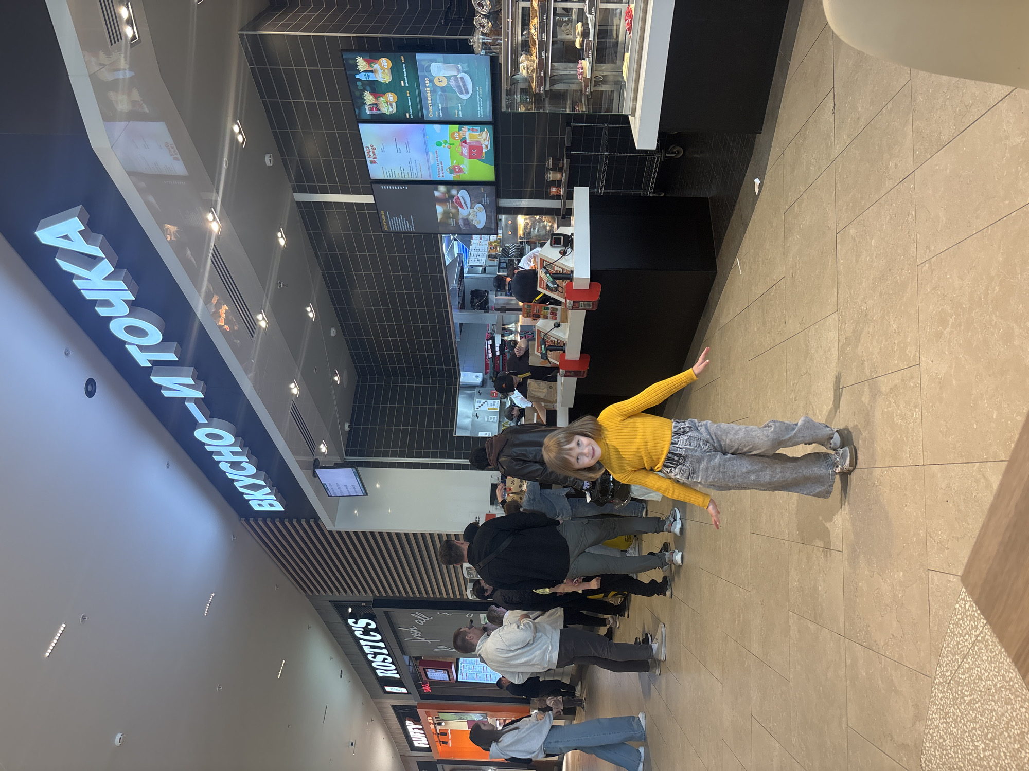 This image captures the interior of a food court in Perm, Russia, specifically focusing on a "ВКУСНО-И ТОЧКА" (Vkusno i Tochka) fast-food establishment and a queue of patrons.
In the foreground, a young girl, approximately 5-7 years old, with light brown hair and a smiling expression, stands facing the camera. She is wearing a bright yellow long-sleeved ribbed top, light wash wide-leg jeans, and light-colored sneakers with pink accents. Her arms are slightly outstretched to her sides.
Behind the girl, a line of approximately eight to ten individuals, mostly adults and at least one child, are queued in front of the "ВКУСНО-И ТОЧКА" counter. Patrons are dressed casually in hoodies, jackets, jeans, and sneakers. The man at the front of the queue wears a grey hoodie and dark pants. Another man in a black jacket and grey pants stands further back in the line.
The "ВКУСНО-И ТОЧКА" restaurant occupies the right side of the frame. Its name, "ВКУСНО-И ТОЧКА," is displayed in large, illuminated blue-and-white letters above the service area. The counter area features a dark tiled wall background. Above the counter, three large digital menu screens display various food items, prices, and promotions, including "Кидз Комбо" (Kids Combo) and "Счастливый час" (Happy Hour). To the right of the counter, a glass display case showcases assorted desserts and pastries. Several restaurant staff members in dark uniforms and visors are visible working behind the counter.
Further down a corridor to the left, another fast-food establishment, "ROSTIC'S," is visible with its name in white text on a black sign. Adjacent to "ROSTIC'S" on the far left, a sign for "FLUFFY" is also visible. Between "ROSTIC'S" and "ВКУСНО-И ТОЧКА," a vertical wall section with light-colored wooden slats provides a decorative element.
The flooring throughout the food court is composed of large, light beige-colored square tiles with dark grout lines. The ceiling is white, with multiple linear and circular recessed light fixtures and smoke detectors/sprinklers. A small, light-colored table corner is partially visible in the bottom right. A metal rolling cart is positioned near the "ВКУСНО-И ТОЧКА" counter. A small piece of white paper litter is visible on the floor near the base of the counter.