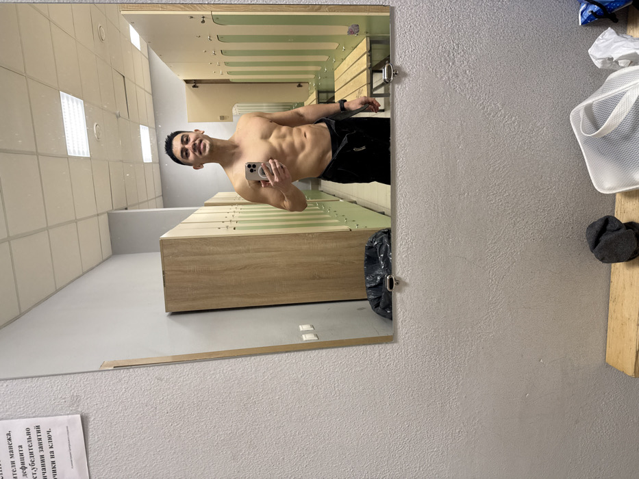 The image displays a male subject taking a mirror selfie in a locker room, identified as being within a sports arena or complex (manege) in Kazan, Russia, based on Cyrillic text visible on a sign.
**People:** One male subject, appearing to be in his mid-20s to early 30s, is captured in a waist-up reflection. He has short, dark hair, and a visible muscular physique with defined abdominal muscles. He is shirtless, wearing dark-colored pants, possibly sweatpants, that are slightly lowered at the waist. A black wristband is on his left wrist. He holds a silver-colored smartphone (likely an iPhone with a MagSafe accessory) in his right hand, extended to take the photograph, and is smiling towards his reflection.
**Subjects/Objects (reflected in mirror):** The locker room features two distinct types of lockers: tall, light-brown wooden lockers on the left, and a row of smaller, pale green compartment lockers with wooden trim and interior hooks on the right. One of the green locker doors is slightly ajar. A light-colored wooden bench runs along the wall opposite the green lockers. A dark cylindrical trash can with a black liner sits on the floor beneath the left row of lockers. The walls are light gray. The ceiling is composed of white acoustic tiles with multiple rectangular fluorescent light fixtures providing illumination. A white sign with black Cyrillic text, visible on the upper left wall, reads "Уважаемые посетители манежа, В случае дефицита просим убедительно сдавать ценные вещи на ключ." (Dear visitors of the arena, In case of a shortage, we kindly ask you to hand in valuable items for safekeeping on key.)
**Subjects/Objects (foreground, not reflected):** The immediate foreground shows a light gray, textured wall. Below the mirror, a light-colored wooden shelf holds a crumpled dark gray sock, a white mesh drawstring bag, crumpled white paper, and a partially visible blue object. Two silver-colored metal fasteners secure the bottom edge of the mirror.
**Actions/Interactions:** The male subject is actively engaging in a mirror selfie, positioning himself to capture his reflection and smiling.
**Environment:** The scene takes place in a clean, brightly lit indoor changing or locker room facility, specifically identified as part of a sports arena or complex (manege) located in Kazan, Russia, by the informational sign.