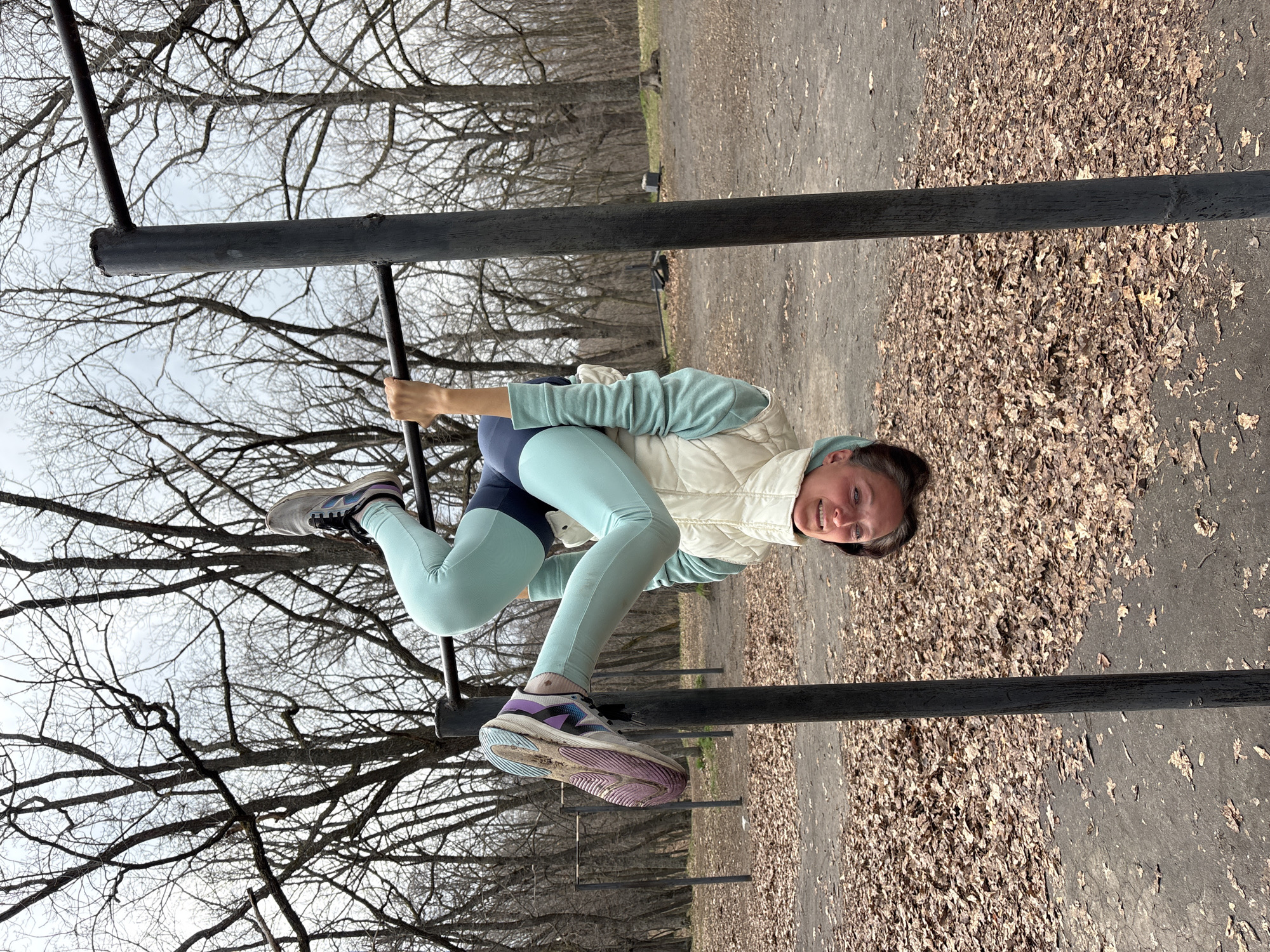 A woman balances on a metal playground structure. She wears athletic leggings, a light vest, and running shoes. The background shows bare trees and a ground covered in fallen leaves. The setting appears to be a park in autumn. The image provides no evidence to indicate the location is Ulyanovsk, Russia.
