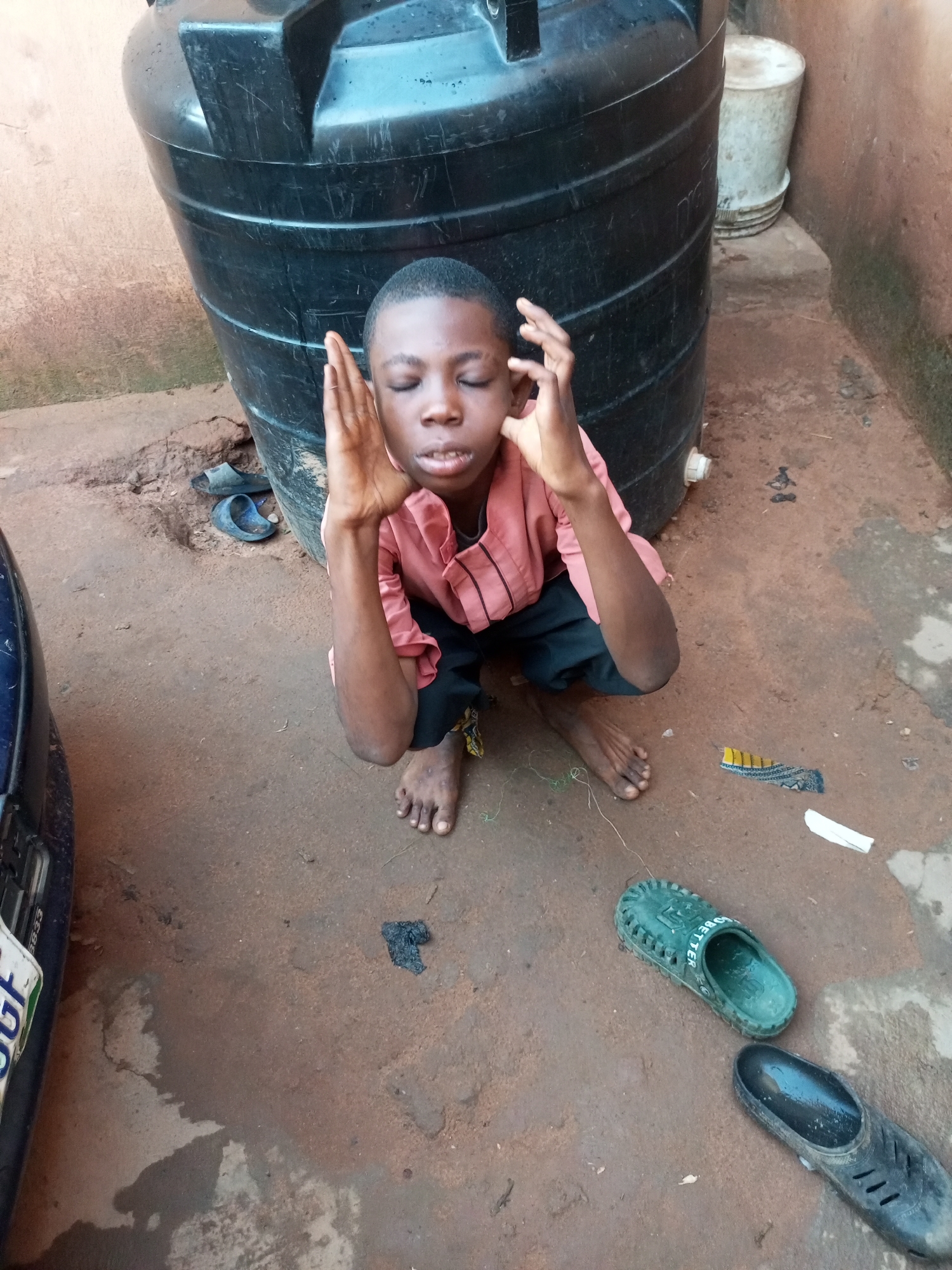 In Tempure, Nigeria, a young person kneels on a dirt and concrete surface near a large black water tank. The individual, dressed in a pink shirt and black shorts, has their eyes closed and hands raised to their temples. Two pairs of sandals are visible on the ground, one blue and one green, along with other debris. A portion of a blue vehicle is to the left of the frame. The environment appears to be an outdoor, utilitarian area, possibly a backyard or storage space, with a light brown wall in the background.