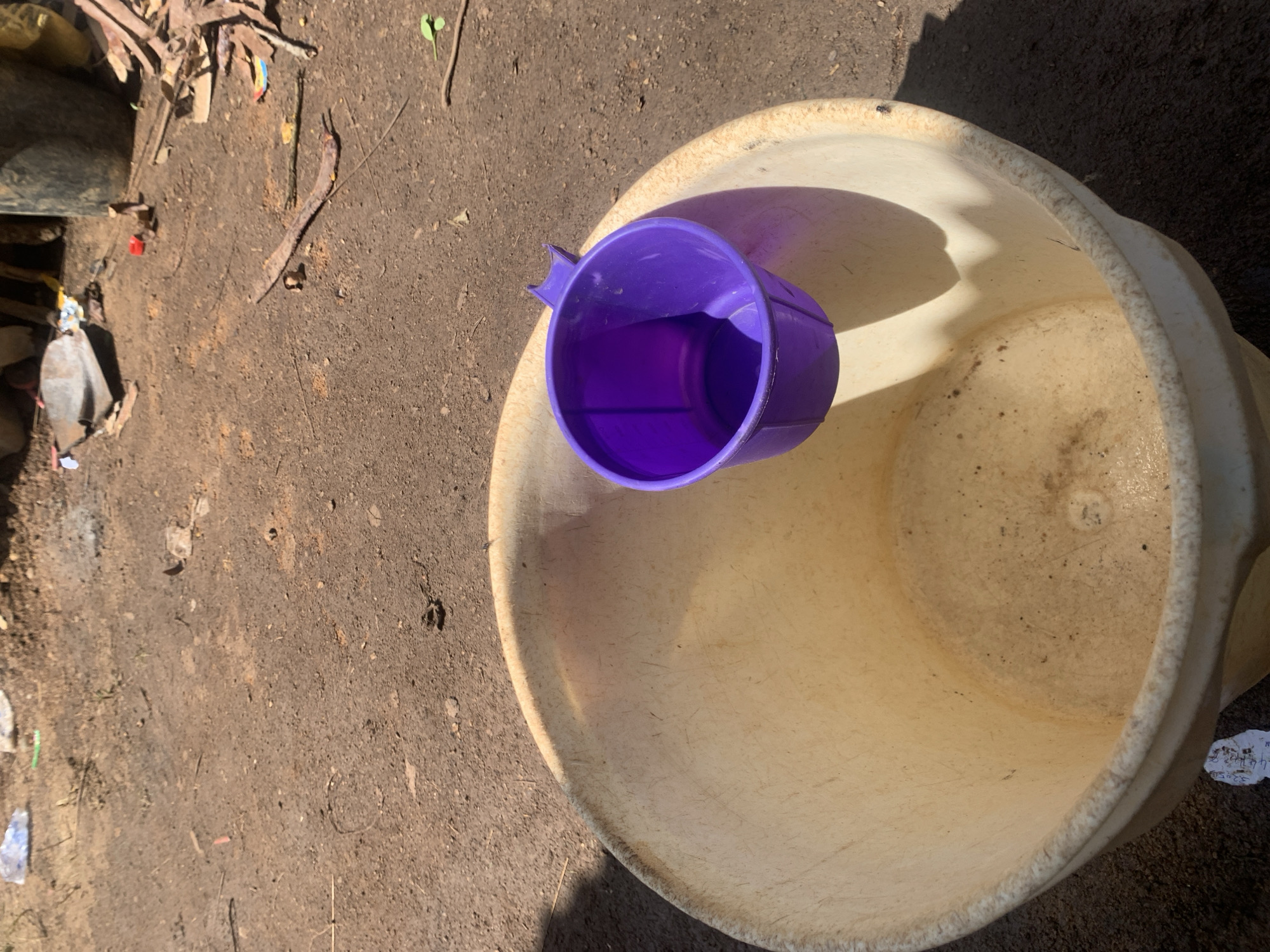 In Buga da Gindi, Nigeria, an outdoor scene depicts a large, worn, cream-colored basin positioned on dry, brown earth. The basin's interior shows signs of use and is slightly grimy. A small, vibrant purple plastic cup rests on the rim of the basin. The surrounding ground is cluttered with various debris, including scraps of paper, plastic, and what appear to be broken pieces of wood and metal. A shovel and a large, dark cylindrical object, possibly another container, are visible in the background. The lighting suggests it is daytime, with clear shadows cast by the objects.