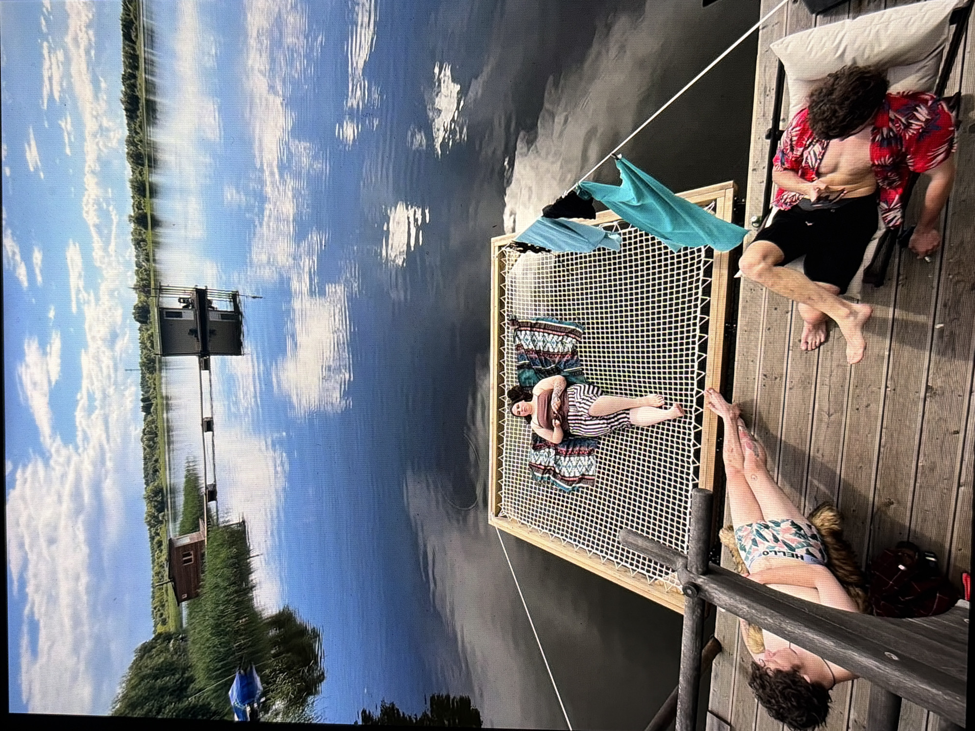 The image shows a serene lake scene, bathed in natural light. Three individuals are visible. One person lies on a net hammock suspended over the water, draped in a striped blanket. Two other individuals are relaxing on a wooden dock in the foreground. One is lying on their side, wearing patterned swim trunks, while the other is reclining with a pillow, dressed in a red floral shirt. The lake reflects the cloudy sky, and a small wooden structure with a pier is visible in the distance. The location is not identified as being in Moscow, Russia.