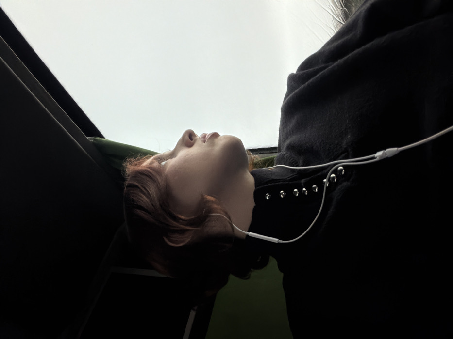 An individual with reddish-brown, wavy hair is observed in profile, seated inside a vehicle in Moineville, France, looking out a window to the right. The person wears a dark, high-collared garment with a vertical line of metallic stud-like fasteners on the left side of the collar. White wired earbuds are connected to the person's left ear, with the wire extending downward over the clothing. A silver hoop earring is visible on the left ear. The upper left portion of the frame shows the dark interior ceiling of the vehicle, with a green fabric element visible on the far left. The window on the right reveals an overcast, light grey sky, with visible rain streaks or condensation on the glass surface. The individual's gaze is directed upwards and outwards, towards the exterior view.