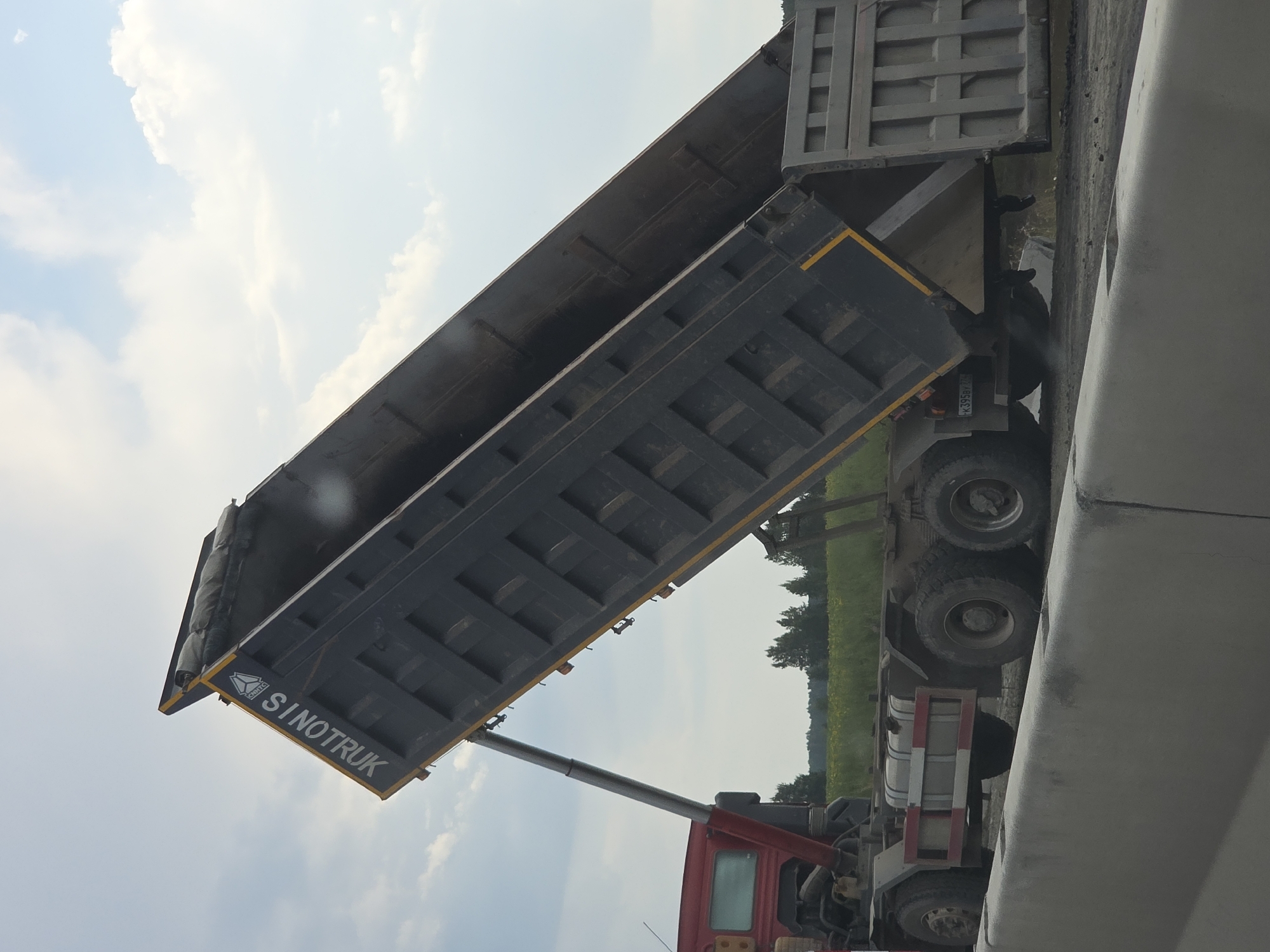 A red and black Sinotruk dump truck is in motion on a paved surface, partially obscured by a concrete barrier in the foreground. The truck's large, dark gray dump bed is raised at an angle, indicating it is in the process of unloading material. The sky above is partly cloudy with a bright, hazy sun. In the background, a flat, green field with scattered trees stretches towards the horizon. The truck's license plate is visible and reads "K395BY 274". The overall scene suggests a construction or industrial setting.