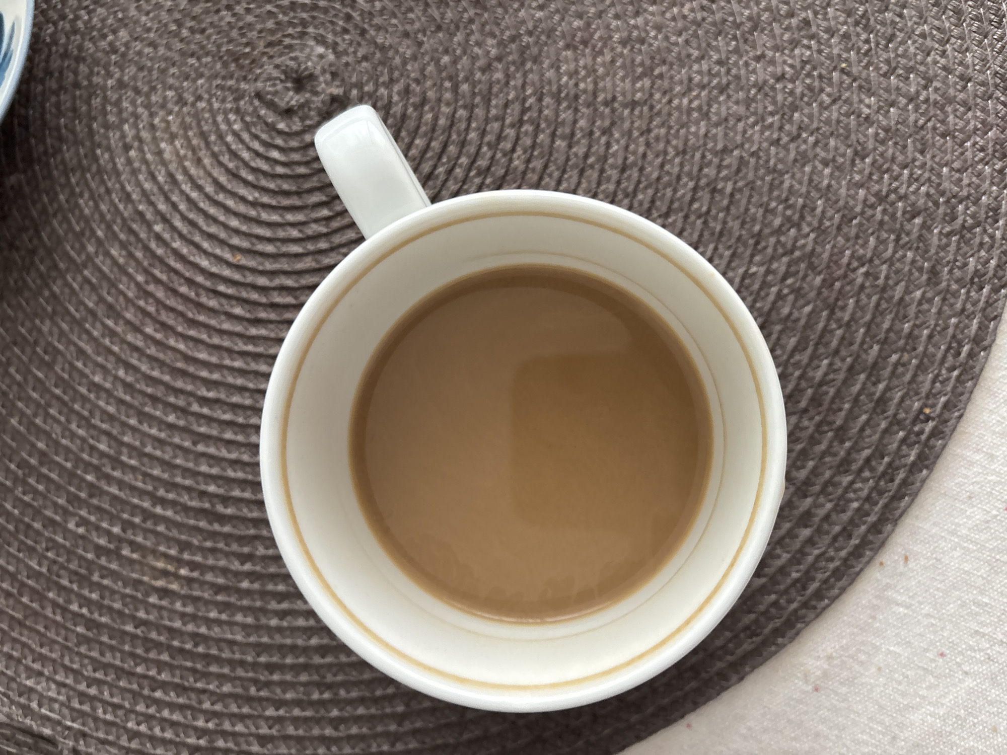 A high-angle, close-up shot of a white ceramic mug filled with a light brown beverage, likely coffee with milk or creamer. The mug has a white handle and a thin gold trim around the inner rim. It sits on a textured, brown circular placemat. Part of a white cloth or napkin is visible on the right side of the placemat. The background is a mix of the placemat and the white cloth. There is no indication of Saint Petersburg, Russia.