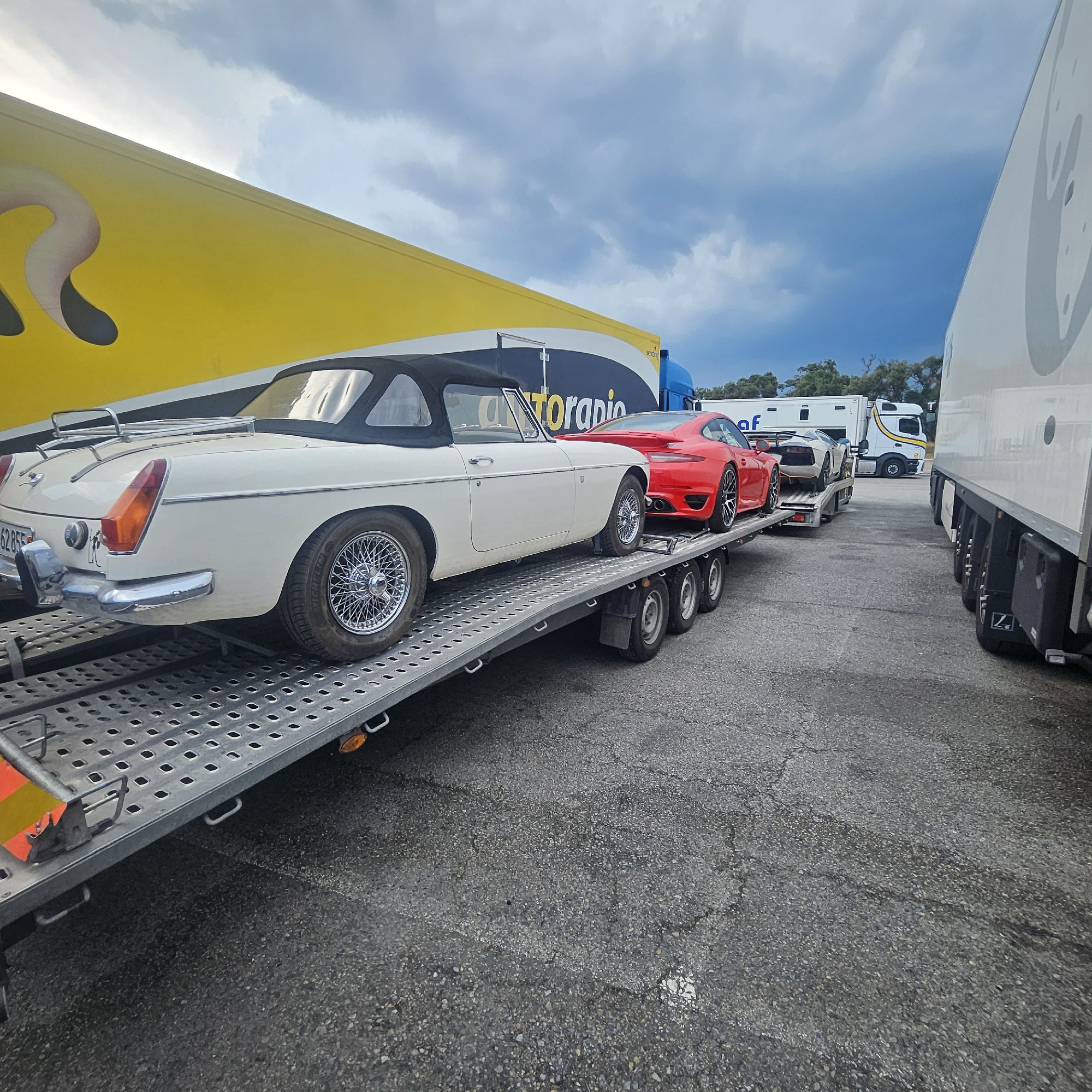 In La Jonquera, Spain, a car transport trailer is parked on a grey asphalt surface. The trailer carries three vehicles: a white convertible classic car, a red sports car, and a white supercar. The trailer is connected to a white semi-truck. A large yellow truck with a logo partially visible is parked to the left of the trailer. The sky is overcast with dramatic clouds.