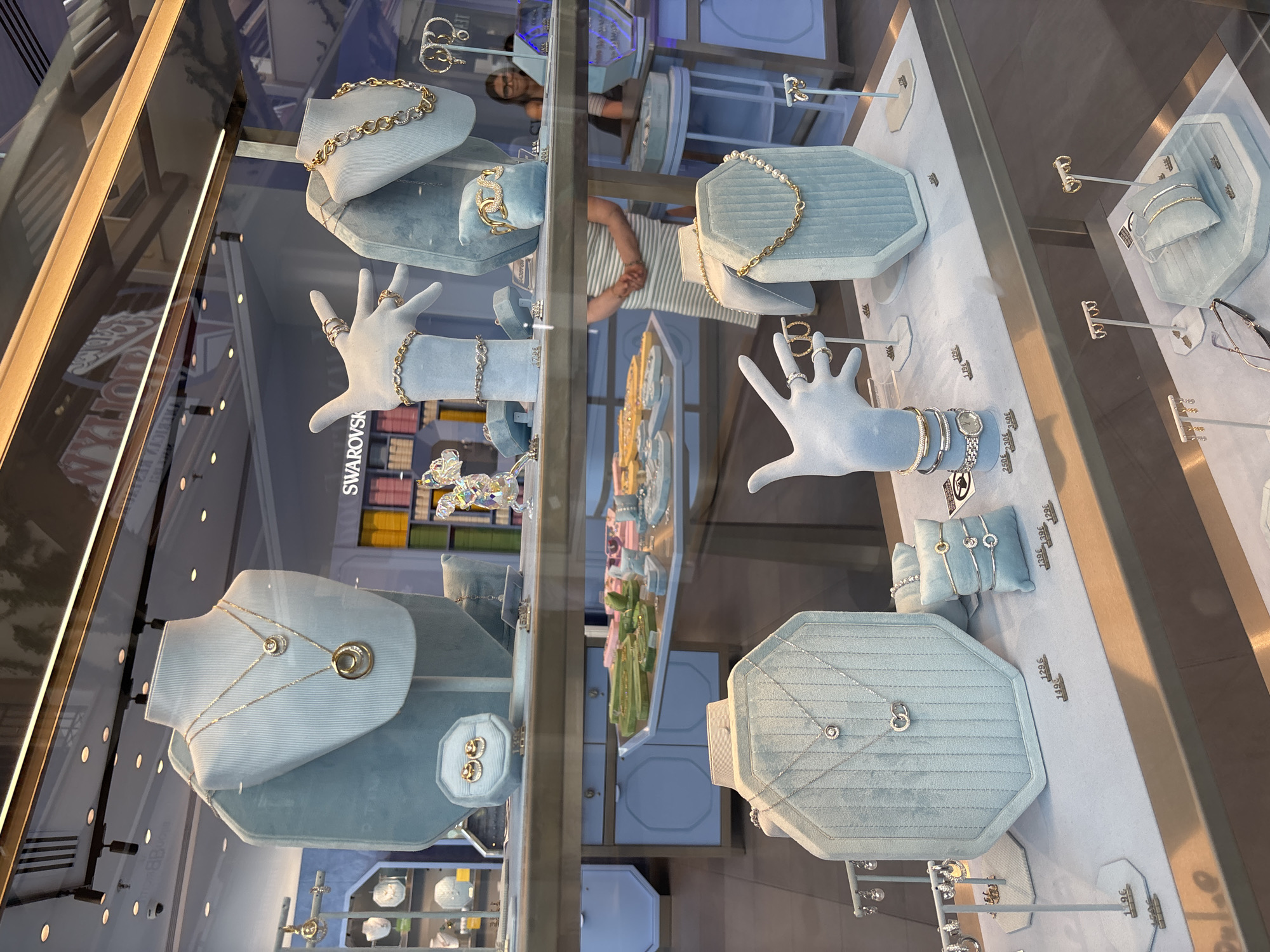 A jewelry store window display is shown, featuring various necklaces, bracelets, and earrings. The display is organized on light blue velvet stands and trays. In the background, a woman with dark hair and glasses is visible, appearing to be an employee. The overall environment suggests a retail setting. The image does not provide any information about the city of Orihuela, Spain.