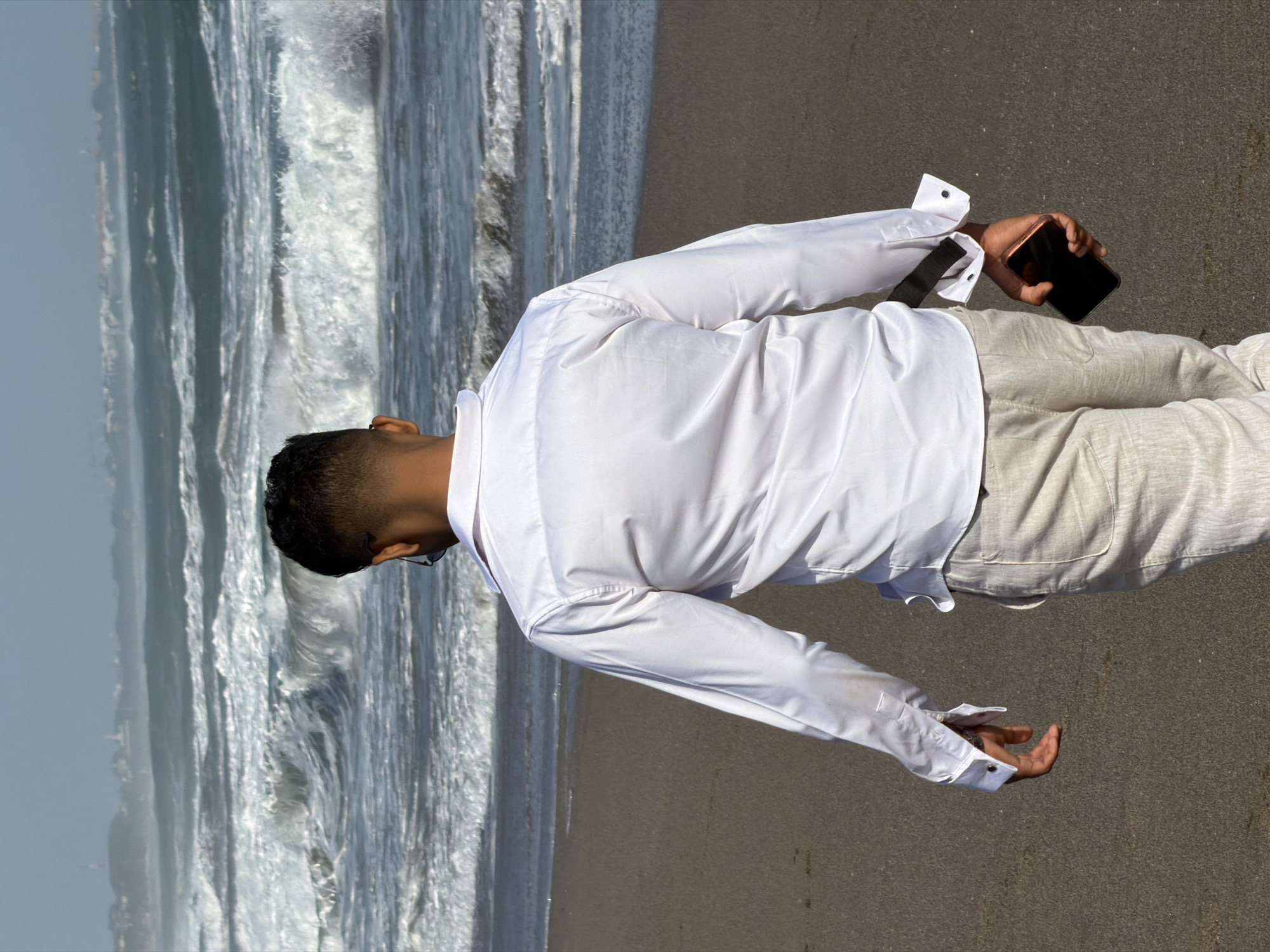A man in a white long-sleeved button-down shirt and beige linen pants walks away from the camera on a dark sand beach. He is holding a black smartphone in his right hand. Behind him, the ocean has large, foamy waves, and a hazy, distant coastline is visible. The scene appears to be the coast of North Kuta, Indonesia.