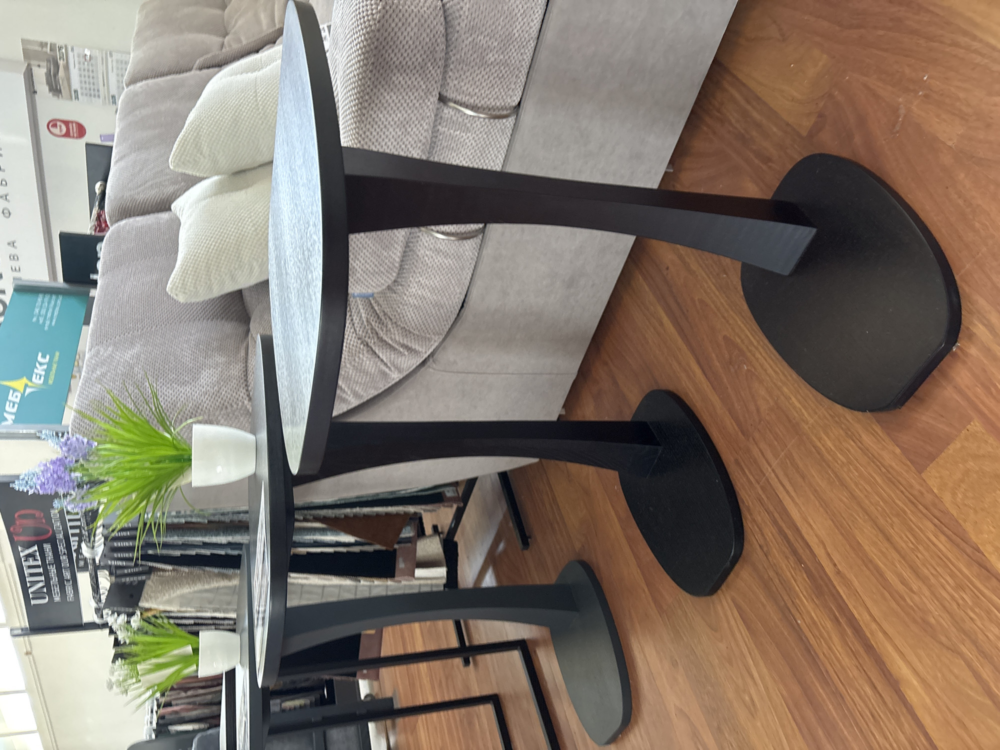 The image depicts an indoor scene, likely a furniture showroom, with several contemporary dark wood accent tables placed on a polished wooden floor. The tables have a distinctive curved pedestal design with oval bases. In the background, a plush sofa with textured upholstery and decorative cushions is visible. To the left of the tables, there's a display rack holding fabric samples and promotional signage, including a "UNITEX" logo and text in Cyrillic. Two potted artificial plants, one with green grass-like foliage and the other with purple flowers, are placed on two of the tables. The lighting suggests an interior setting with artificial light sources. There are no people or animals present in the frame. The scene does not provide any specific indicators for the city of Zaporizhzhia, Ukraine.