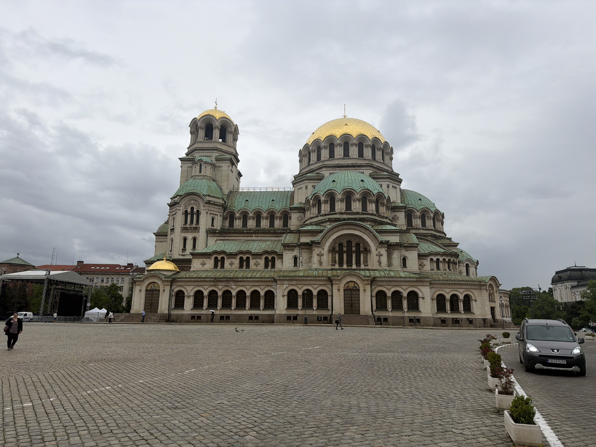 The image shows the Alexander Nevsky Cathedral in Sofia, Bulgaria, on a cloudy day. The cathedral's architecture is prominent, featuring golden domes and green roofs. In the foreground, there is a large cobblestone square, with a few people walking across it. To the right, a gray car is parked near potted plants lining the edge of the square. A stage or structure is set up on the left side of the frame, and other buildings are visible in the background. The overall scene is a typical urban landscape with the iconic cathedral as the focal point.