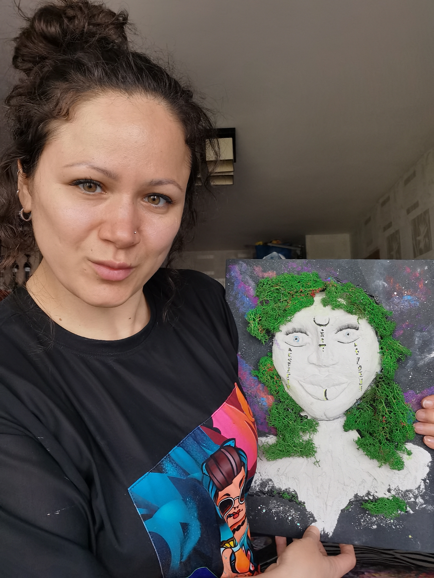 A woman with curly dark hair tied in a bun, brown eyes, and a nose piercing, wearing a black t-shirt with a colorful graphic, holds up a piece of artwork. She is looking directly at the camera with a subtle expression.
The artwork is a 3D relief sculpture of a face mounted on a canvas, appearing to be crafted from plaster or a similar white material. Green moss is used to represent the hair and parts of the chest or shoulders, giving it a natural, organic look. The face features drawn eyebrows, elongated blue eyes, and symbols or glyphs drawn on the forehead (including a crescent moon shape and vertical lines) and along the cheeks/jawline. The background of the canvas is dark with splatters of purple, blue, and pink, resembling a galaxy or cosmic scene. Below the sculpted face, the white material extends downwards, suggesting a bust or flowing garment.
The setting appears to be indoors, likely a home, with a plain ceiling and a wall with textured wallpaper visible in the background. The lighting is bright and natural, suggesting daytime. The woman holds the artwork with both hands, presenting it. The location is Chaikovsky, Russia.