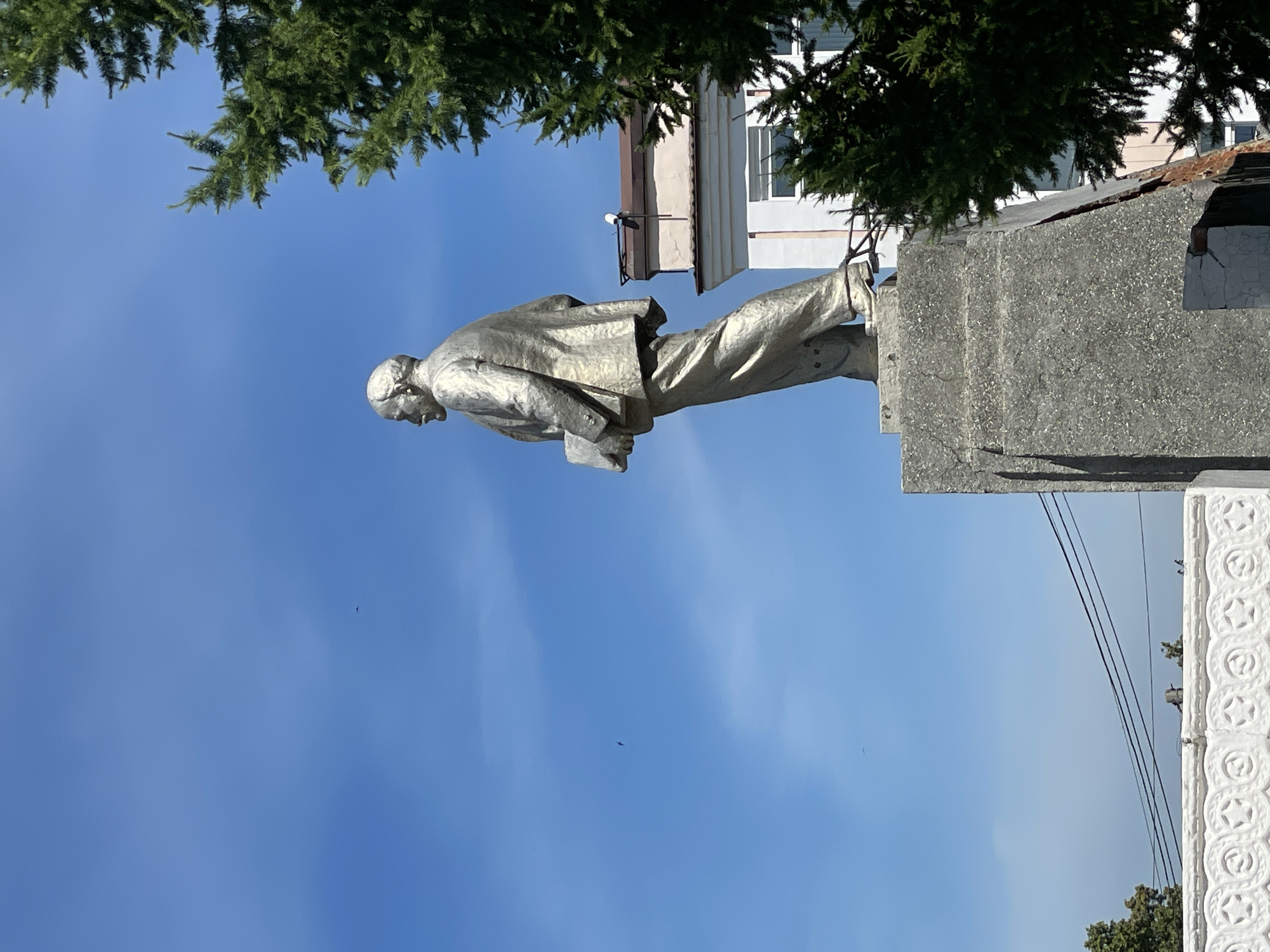 A full-body silver statue of Vladimir Lenin stands on a concrete pedestal, facing left. Lenin is depicted wearing a suit and holding a book in his left arm. The statue is positioned in an outdoor urban environment under a clear blue sky. A large green coniferous tree is visible on the right side of the image, partially obscuring a building in the background. The building has a light-colored facade with windows and a tiled roof. There are also power lines visible across the scene. The base of the pedestal shows some signs of wear and damage.
