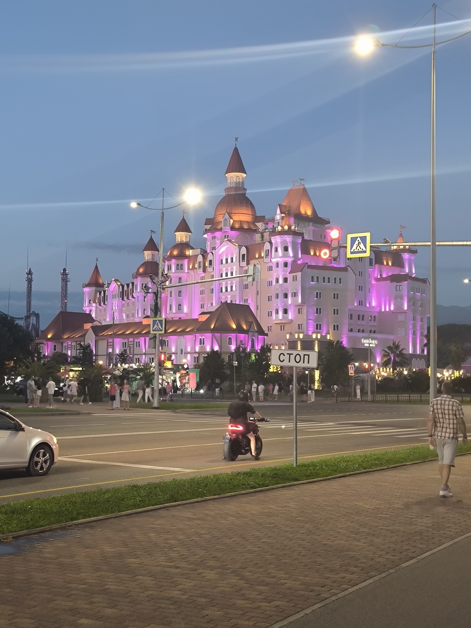 The image depicts a twilight scene in Sirius, Russia, featuring the brightly illuminated Radisson Collection Paradise Resort & Spa. The resort, with its pink and orange hues, dominates the background, its fairy-tale-like architecture complete with turrets and illuminated windows. In the foreground, a paved walkway borders a strip of green grass and a street. A silver car is visible on the left side of the street, and a person on a black motorcycle is moving across the crosswalk on the right. Further down the street, a person in a plaid shirt and shorts is walking away from the camera. Pedestrians are milling about on the sidewalk in front of the resort, and traffic lights and street lamps are visible. The sky is a deep blue, indicating the transition from day to night.