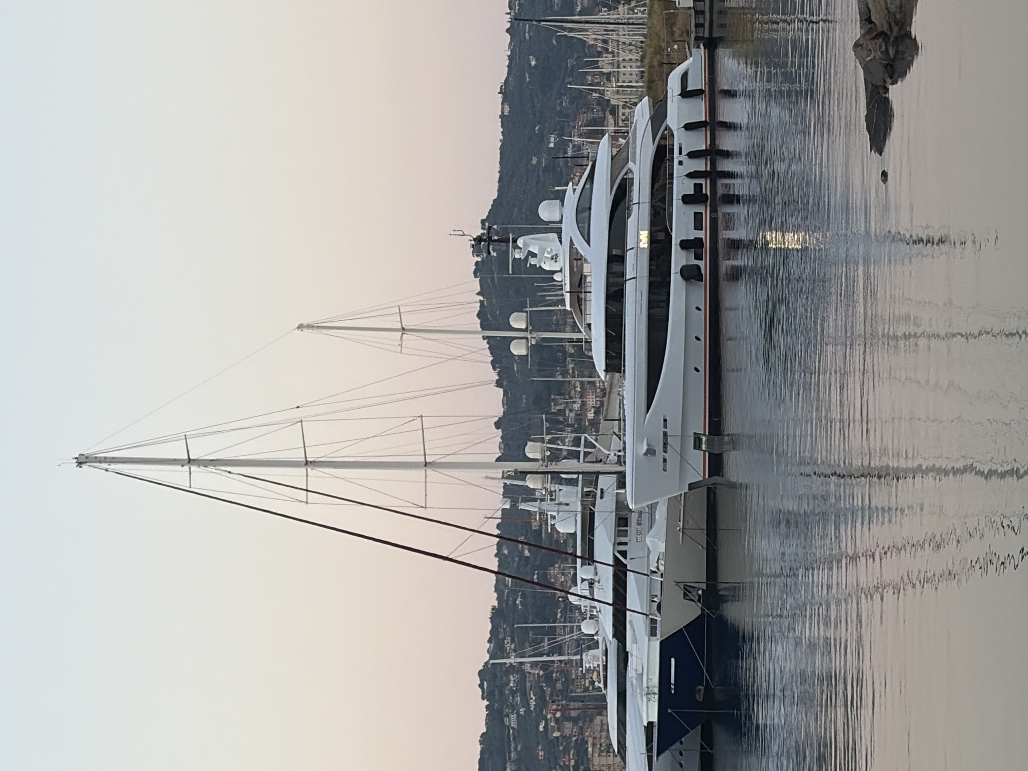 The image depicts a fleet of luxury yachts docked in a harbor, with the town of Imperia, Italy, visible in the background. The yachts are white with dark trim and red accents. The masts of sailboats are also prominent in the foreground and background, suggesting a marina catering to various types of vessels.
The setting is a waterfront with calm water reflecting the sky and the boats. A rocky outcrop is visible on the right side of the frame, emerging from the water. The sky is overcast with a soft, diffused light, indicating either early morning or late afternoon. The hills behind the town are covered in lush green vegetation and dotted with buildings.
There is no discernible activity taking place; the yachts appear to be at rest. The overall mood is serene and tranquil, emphasizing the beauty of the harbor and the elegance of the vessels. No visible text is present in the image.