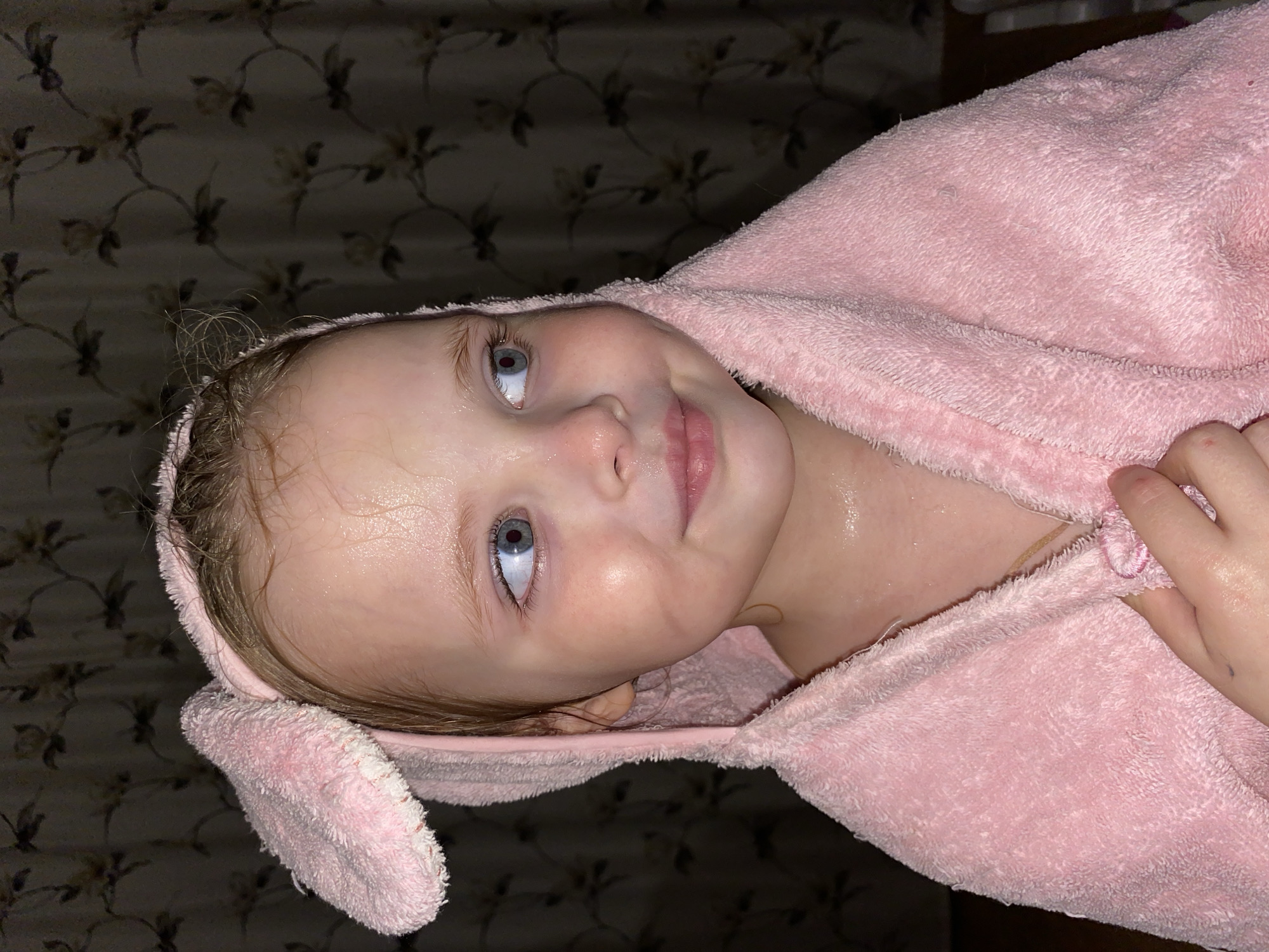 This close-up portrait captures a young child, likely a toddler or preschooler, in an indoor setting in Votkinsk, Russia. The child, with fair skin, light hair, and expressive blue eyes, is wrapped in a soft, pink hooded towel. Their hair and face appear wet and shiny, suggesting they have recently had a bath or shower.
The child's gaze is directed upwards and slightly to the right, showing a moment of curiosity or engagement, possibly with a parent or caregiver. A gentle, faint smile plays on their lips, conveying a sense of contentment or slight amusement.
The background features a dark, patterned wall covering or textile, with an abstract floral or vine motif in muted, earthy tones. The lighting is dim and warm, indicative of artificial indoor lighting, likely setting a cozy atmosphere, possibly in the evening after a bath. No text is visible in the image.