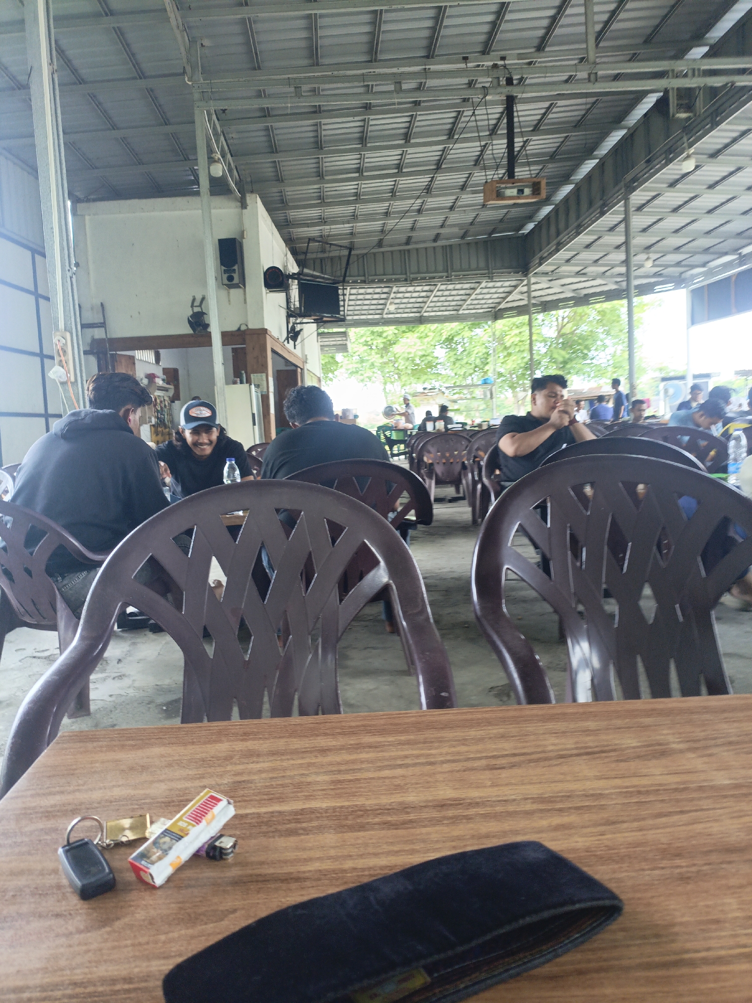The image depicts an outdoor seating area, likely a casual eatery or rest stop, under a large metal roof supported by columns. The atmosphere is relaxed, with several people seated at tables. In the foreground, a wooden table displays a set of keys, a pack of cigarettes, and a black wallet.
In the middle ground, three men are seated at a table. One man, wearing a black hoodie, is facing away from the camera. Another man, in a black t-shirt and a blue baseball cap, is laughing and looking towards the man in the hoodie. A third man, also in a black t-shirt, is partially visible. They are surrounded by dark brown plastic chairs.
In the background, more people are seated at tables, and trees are visible through the open sides of the structure. The overall setting appears to be a communal gathering place. Based on the context provided, this scene is located in the city of Lhokseumawe, Indonesia.