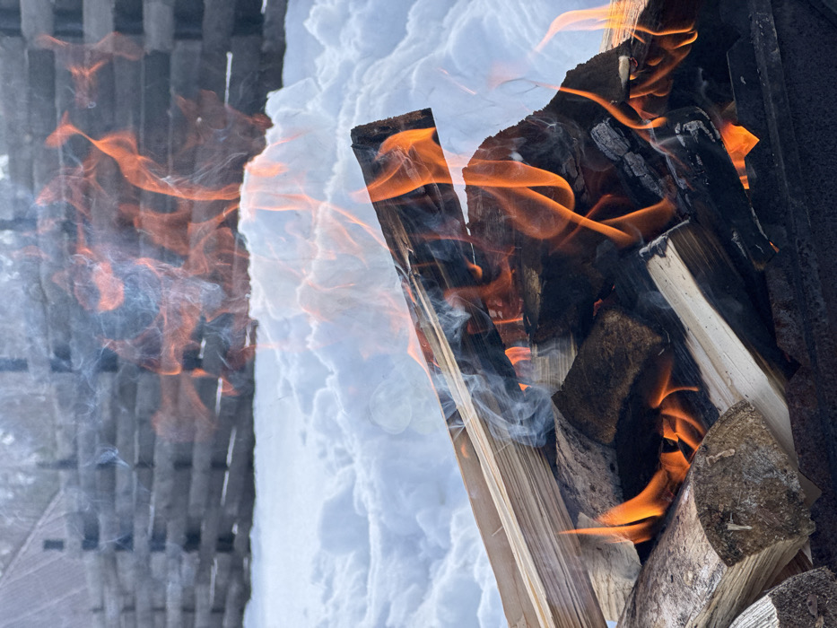 An outdoor scene in Kamskoe, Russia, features an active fire burning split firewood. Bright orange and yellow flames engulf multiple pieces of wood, which vary in color from light tan to dark, charred brown. Columns of grey-white smoke intermingle with the flames, rising from the burning material. The fire appears contained within a dark, possibly metallic, structure at the bottom of the frame. Immediately behind the fire, deep, undisturbed white snow covers the ground, forming uneven drifts. In the upper background, a dark, weathered wooden fence or retaining wall, constructed from horizontal timbers, is visible, partially obscured by the snow and smoke. The sky above is overcast, and no individuals are present.