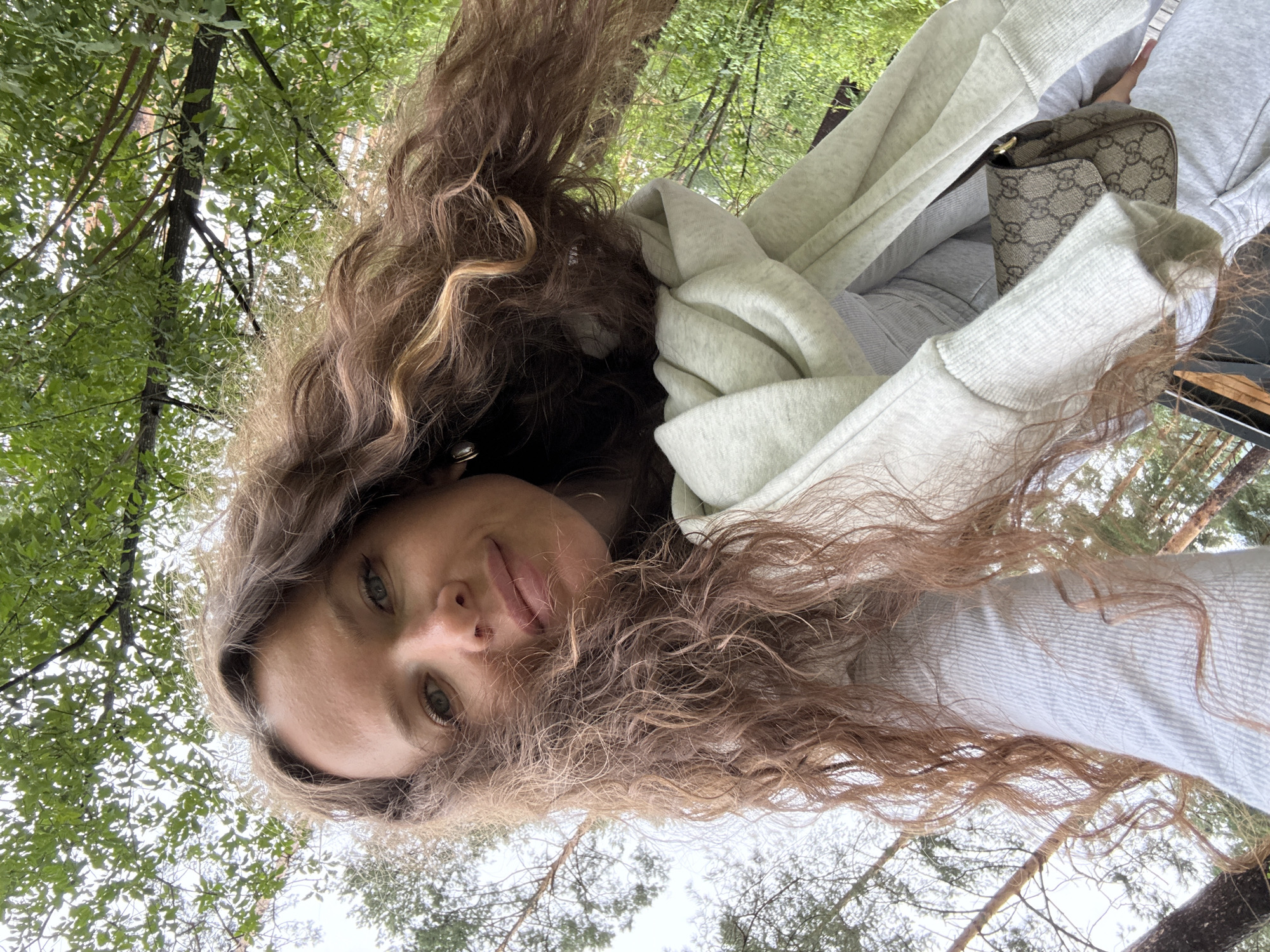 A low-angle, close-up selfie captures a young Caucasian woman with long, curly brown hair and green eyes looking towards the camera. She is wearing a light gray hooded sweatshirt, with a white scarf draped over her shoulder, and a small brown designer handbag with a monogram pattern is visible. The background consists of lush green trees and a cloudy sky, suggesting an outdoor setting likely in the city of Nizhnekamsk, Russia, possibly during the summer or early autumn. The perspective of the shot, taken from below, emphasizes her face and hair against the natural backdrop.