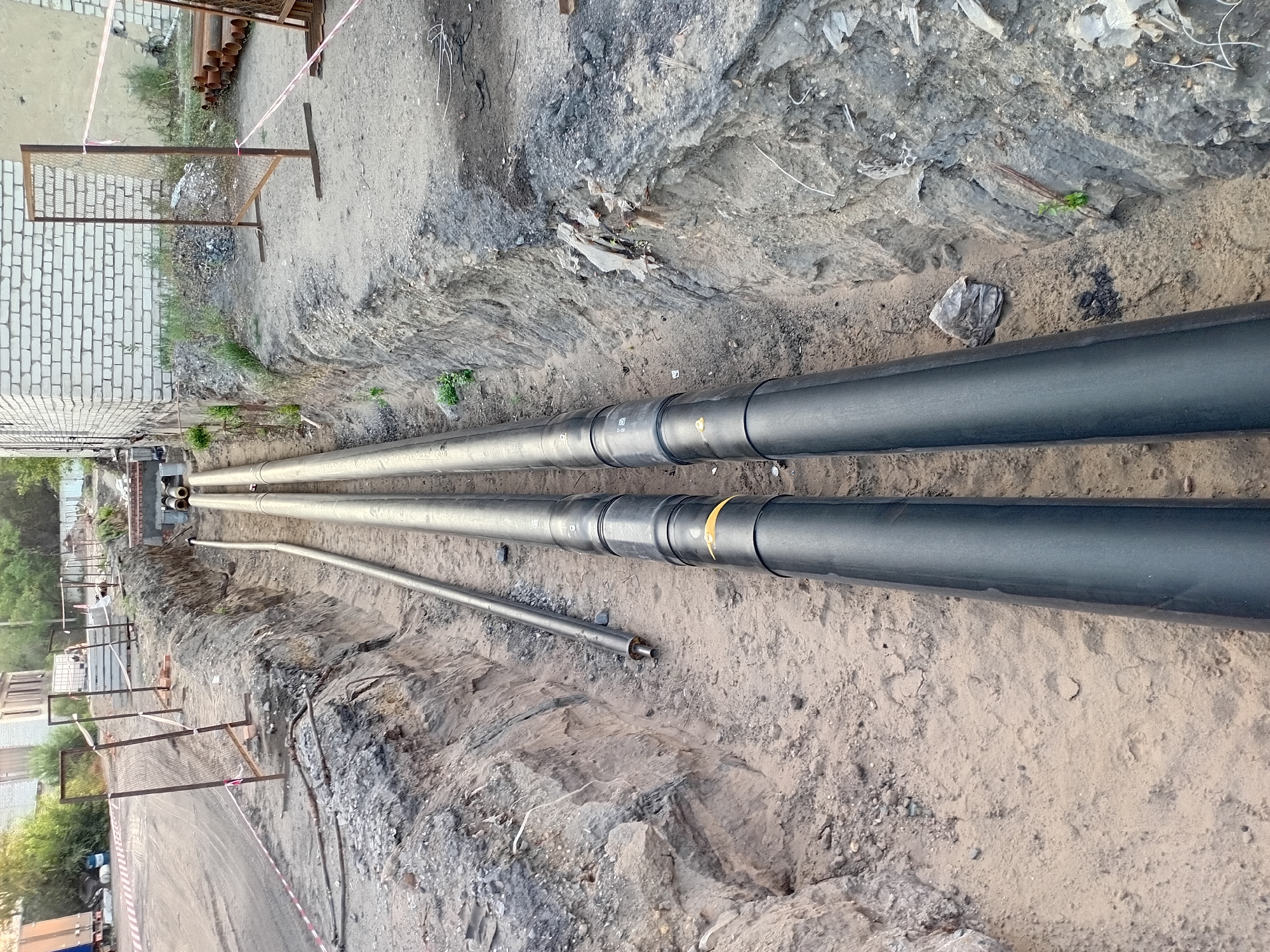 The image depicts an outdoor construction scene, likely in the city of Semey, Kazakhstan, focusing on an excavated trench. Two large, black pipes, possibly for utilities, are laid side-by-side within the trench. A thinner, metallic pipe is positioned to the left of the main pipes. The trench walls are made of soil and gravel, showing signs of recent excavation. Fencing with orange and white caution tape is visible along the edge of the trench, indicating a work zone. In the background, a brick building and stacks of metal pipes are present. The ground appears to be unpaved, consistent with a construction site. There are no people visible in the image, and no specific actions or interactions are depicted. The overall environment suggests ongoing infrastructure work.