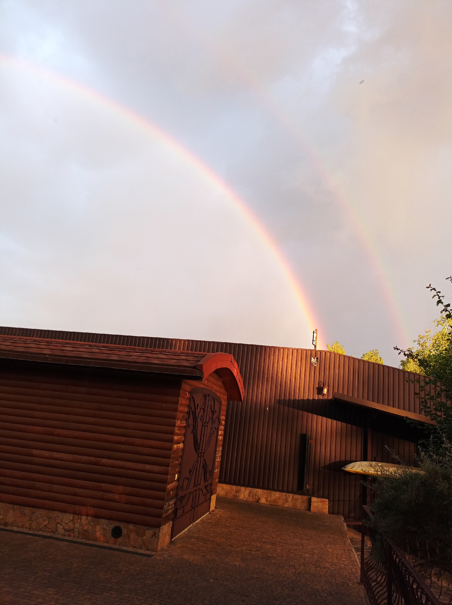 This is a landscape photograph taken in Kozyn, Ukraine, featuring a double rainbow arching across a cloudy sky. In the foreground, a wooden building with a textured, dark brown facade and a red-tiled roof is visible. A large, ornate metal gate is attached to the building. A paved walkway leads away from the building. The scene is illuminated by the warm, golden light of either sunrise or sunset, casting long shadows. To the right of the building, a metal railing and some foliage are present. The overall atmosphere is serene and picturesque, with the natural phenomenon of the rainbow contrasting with the man-made structures.