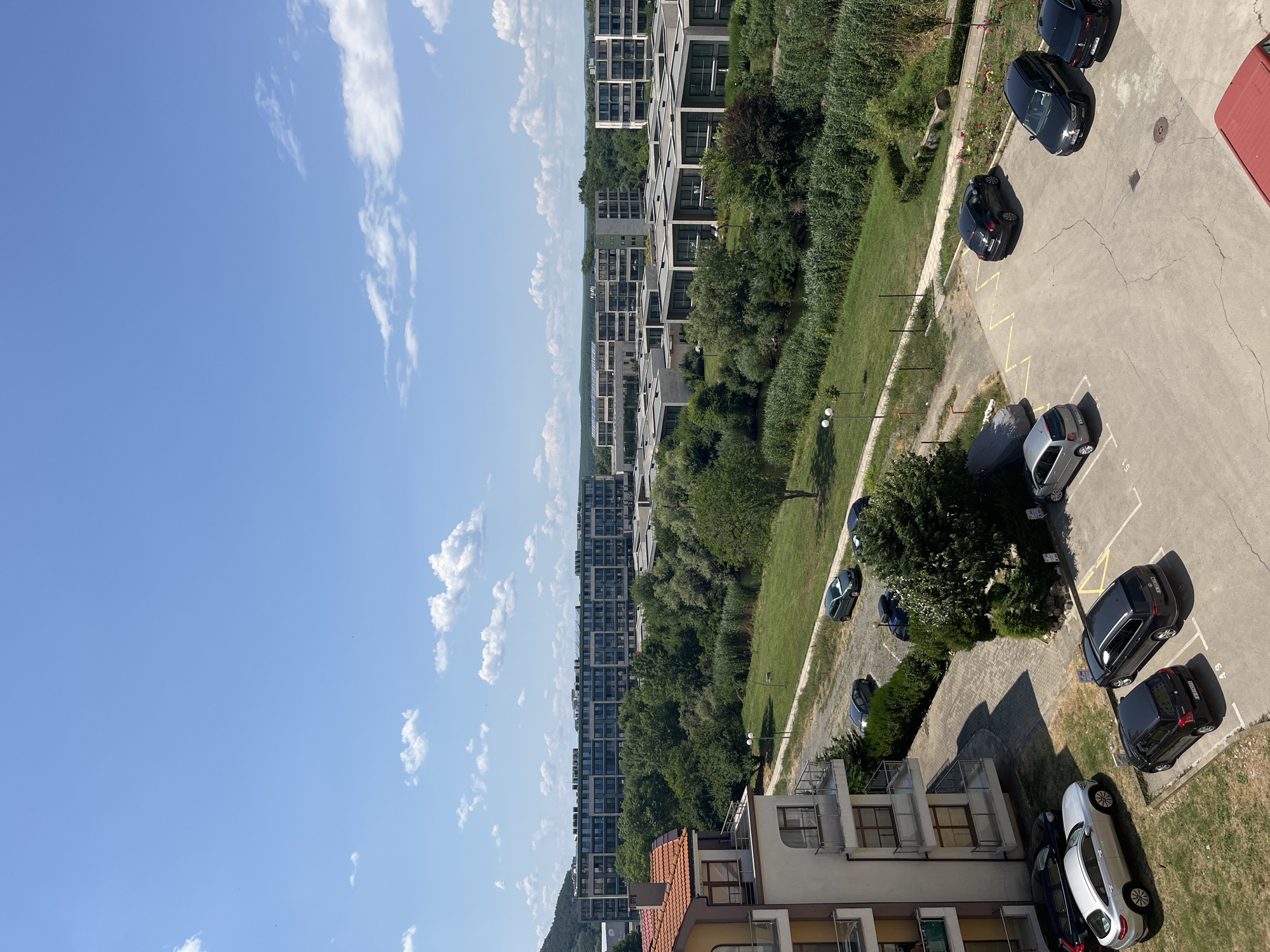 The image captures a wide view of a residential area in Obzor, Bulgaria, on a sunny day with scattered clouds. In the foreground, a building with a red-tiled roof and beige facade is partially visible, featuring balconies. A parking lot is situated in front of this building, with several cars parked. These include a silver Mercedes-Benz, two black sedans, a dark SUV, and a silver hatchback. Further back, a green grassy area is interspersed with trees and dense vegetation. Beyond this, a complex of modern, multi-story apartment buildings with many windows and balconies dominates the middle ground. The architecture appears contemporary, with flat roofs and light-colored facades. In the distance, more buildings and greenery are visible, with a hillside forming the backdrop. The sky is a clear blue, with fluffy white clouds. The scene suggests a well-developed urban or semi-urban environment, likely a resort or residential complex given the proximity to greenery and the style of the buildings.