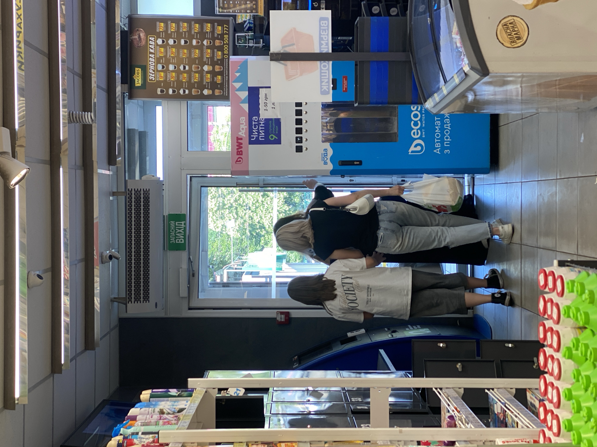The image depicts the interior of a small convenience store or market. In the foreground, a row of green bottles with red caps is visible on a shelf, followed by a display of white bottles with red caps. A bright pink and blue water vending machine is prominent on the right side of the frame, displaying text in Ukrainian, including "BWT Aqua" and "Чиста питна вода" (Clean drinking water). To the left of the vending machine, a blue kiosk-like structure with various compartments is present.
The background shows an open doorway leading outside, with trees and greenery visible. Above the doorway, a green sign with white Ukrainian text reads "ЗАПАСНИЙ ВИХІД" (Emergency exit). A coffee advertisement poster is mounted on the wall to the right of the doorway, featuring various coffee options and prices in Ukrainian. Several security cameras are visible on the ceiling.
Three individuals are in the middle ground, facing away from the camera and walking towards the doorway. The person in the center wears a white t-shirt with "SOCIETY" printed on it in black lettering, paired with grey shorts and black sneakers. The person to their right wears a black t-shirt and light blue jeans, carrying a white shopping bag. The third person, partially obscured, appears to be wearing a dark top and light-colored pants.
The environment suggests a commercial establishment, likely a local store. The presence of specific Ukrainian text and signage indicates the location is in Ukraine. Considering the context of similar images and the typical branding of such establishments, this scene is likely in Poltava, Ukraine.
