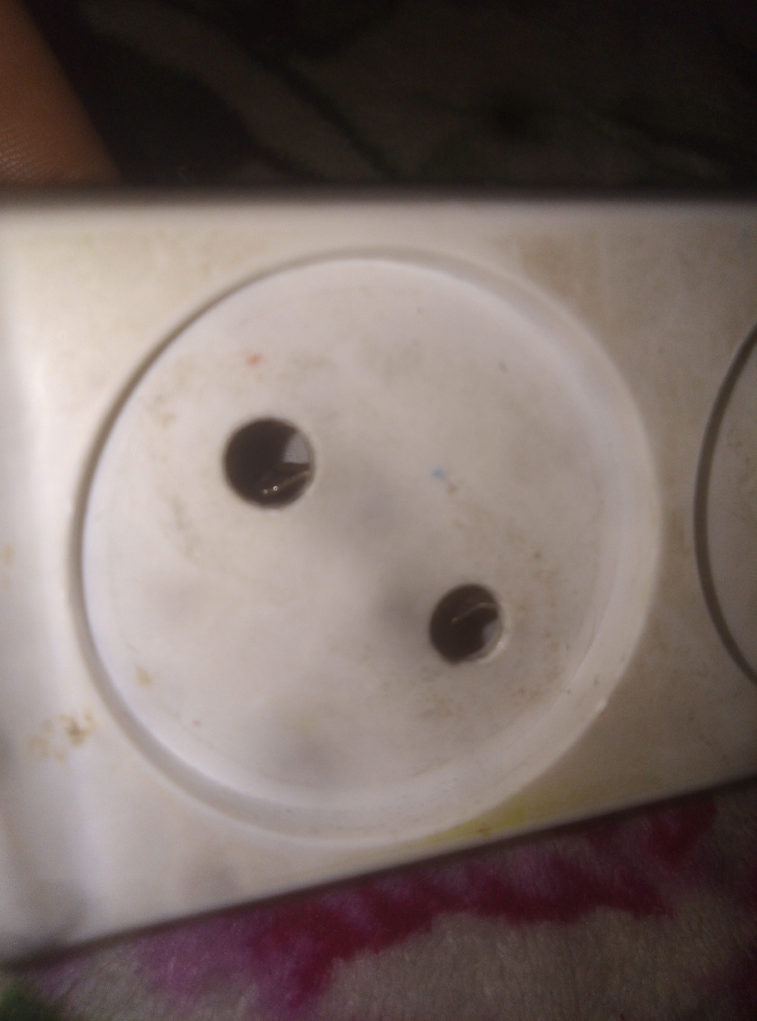 A close-up, slightly blurry photograph depicts an electrical outlet. The outlet is made of off-white or light-colored plastic and features two round receptacles, characteristic of Type E or Type F (Schuko) sockets commonly used in many parts of the world. The surface of the outlet appears aged and somewhat dirty, with visible grime, scuffs, and areas of discoloration, including yellowish tints and small reddish or bluish marks. Inside the left socket hole, metallic electrical contacts are visible. The background is out of focus; the upper portion shows dark, indistinct shapes, while the lower part reveals blurry patches of reddish-purple and brown, possibly indicating a fabric or patterned surface beneath the outlet. No people or activities are depicted, and there is no visible text. The lighting suggests an indoor setting.