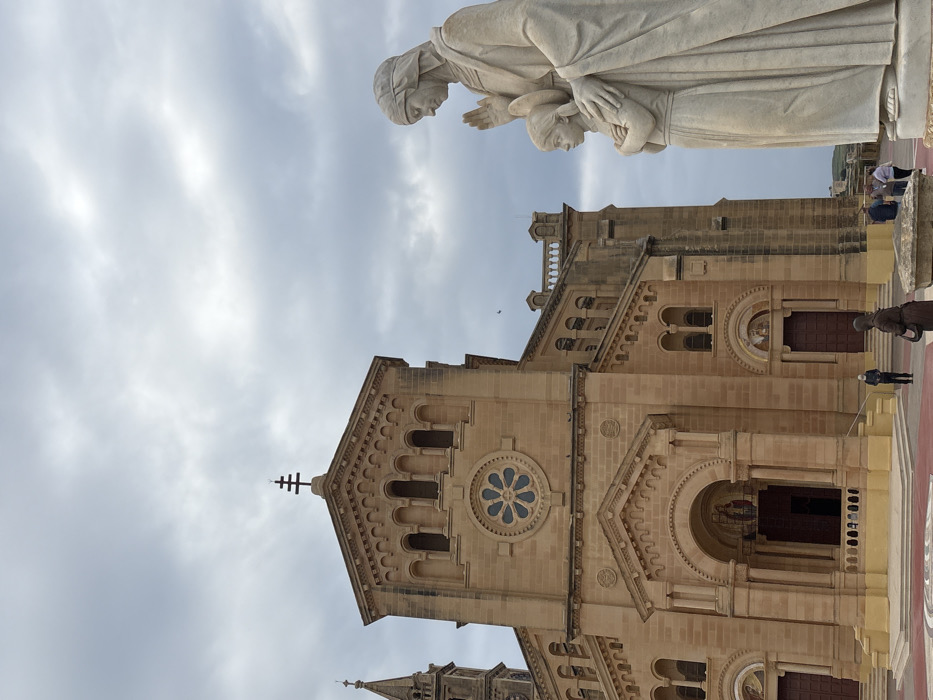 The image depicts the exterior of the Basilica of the National Shrine of the Blessed Virgin of Ta' Pinu, located in Għarb, Gozo, Malta, under an overcast sky. The prominent structure, constructed of light brown limestone, features a central facade with a large arched entrance containing mosaic artwork, topped by a circular rose window. Above this, a triangular pediment is crowned with a unique double-bar cross. A tall bell tower with a pointed spire is partially visible on the far left. To the right, a large light-colored stone statue stands, portraying an adult female figure with her right hand raised and her left hand resting on the shoulder of a smaller female figure, whose arms are crossed. In the foreground, near the church steps, one individual in a dark garment is visible, along with another person in a blue jacket wearing a face mask. Further right, a small group of three to four individuals is gathered. The ground in front of the basilica is paved with geometric patterns in red, brown, and yellow tones. Rolling green landscape is visible in the background on the right.