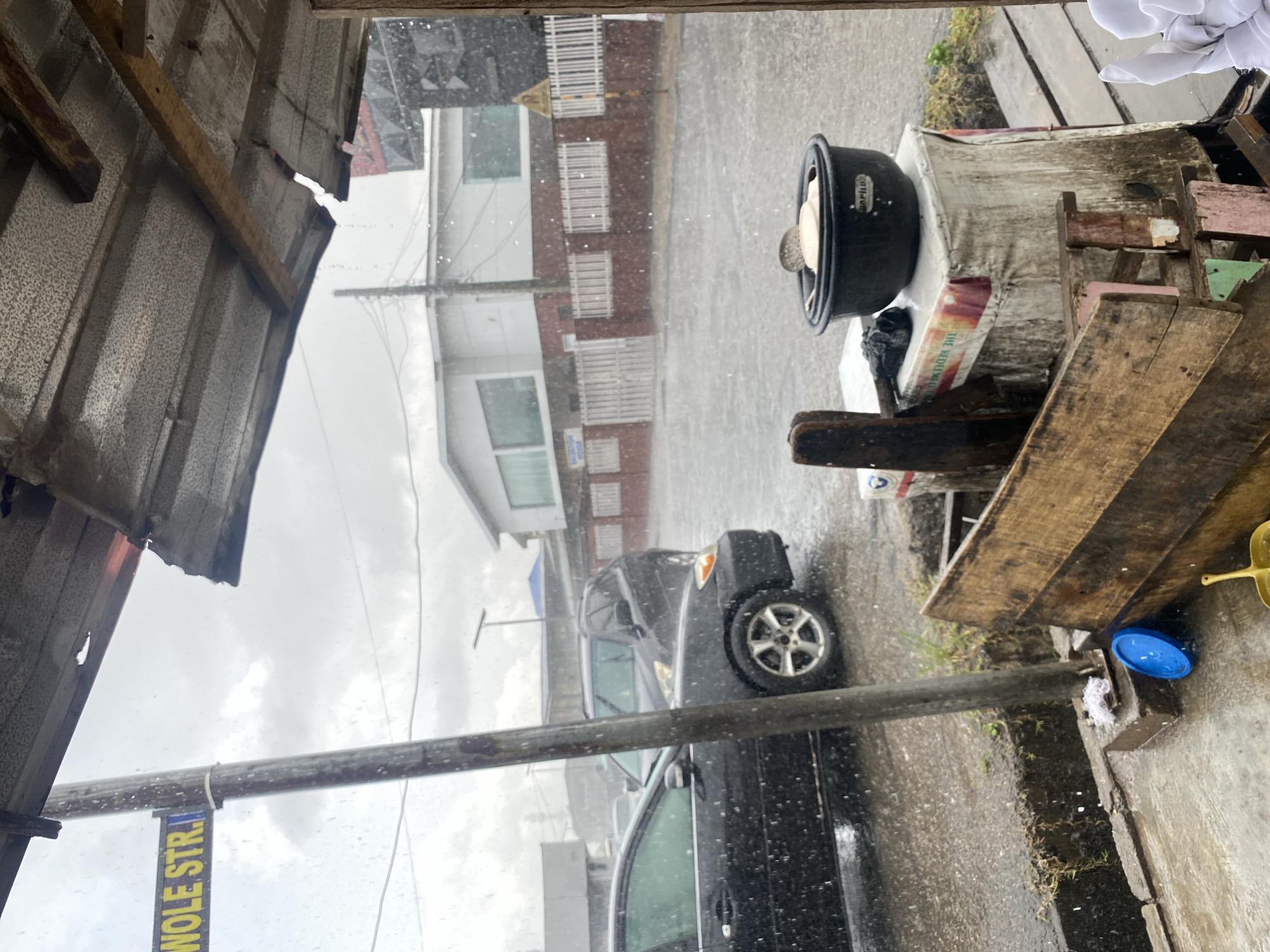 The scene shows a rainy day in Lagos, Nigeria, as indicated by the visible "Wole Street" sign. The view is from under a corrugated metal structure. Outside, rain falls heavily on a street where a black car is parked. A wooden cart with a black pot on top is situated to the right of the frame. The surrounding buildings appear to be residential or commercial spaces with white and brick facades.